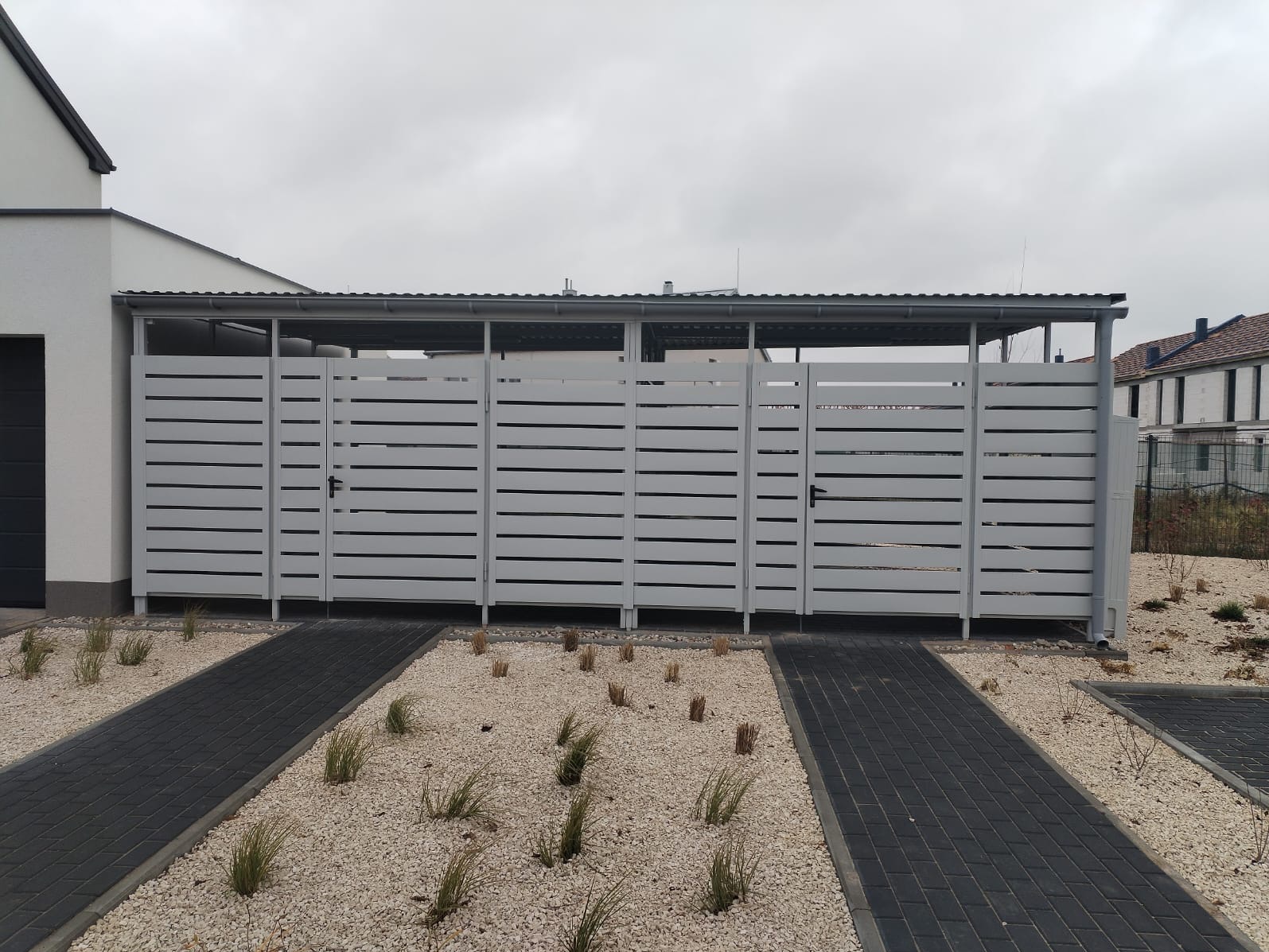 Szary, nowoczesny garaż z blachy z poziomymi panelami, zadaszony, z czarną ścieżką z kostki brukowej i białym żwirem z roślinami.