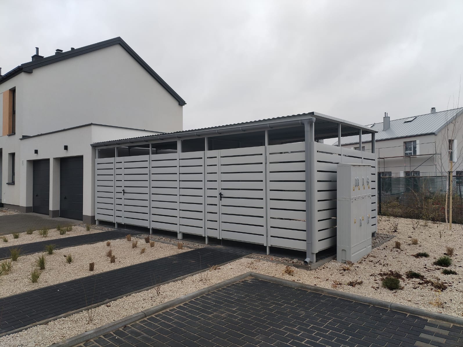 Nowoczesny, szary garaż blaszany z poziomymi panelami, zadaszony, przylegający do domu z dwoma garażami, otoczony żwirem i kostką brukową.