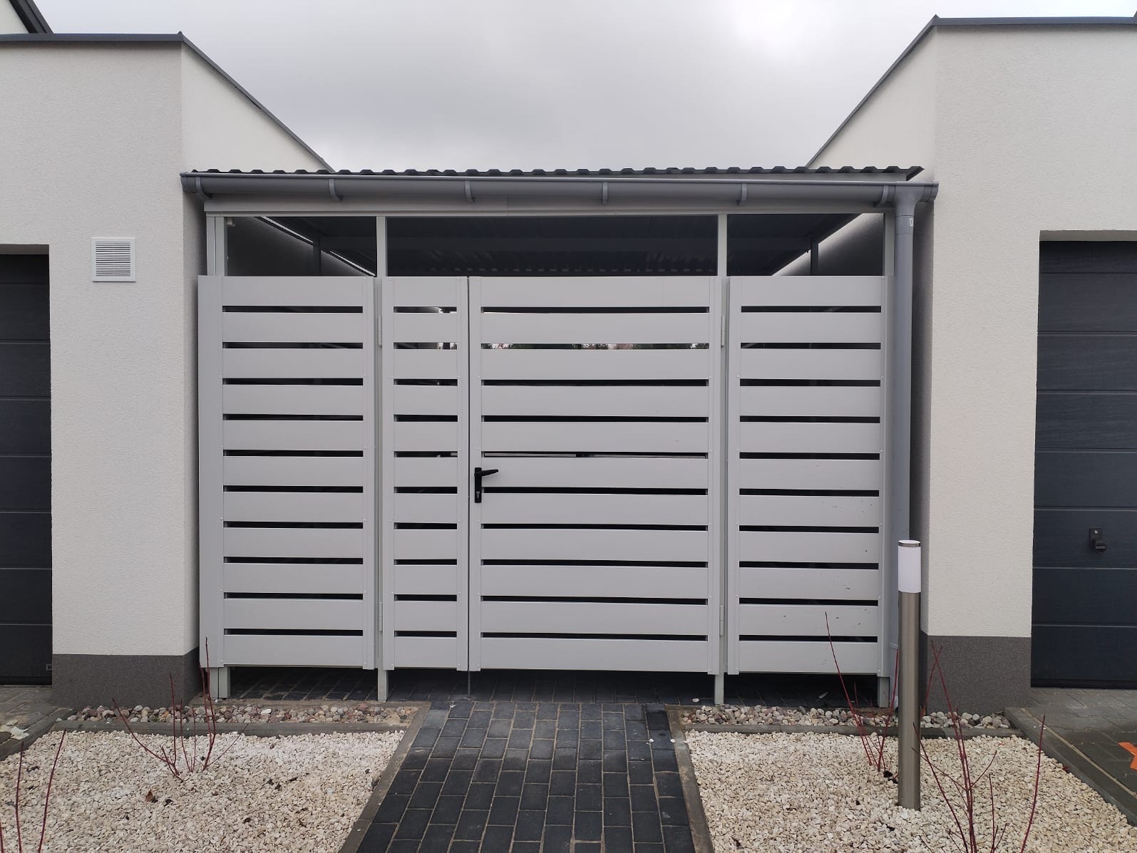 Nowoczesna, biała brama garażowa lamelowa z czarnym uchwytem, osłonięta daszkiem, z oświetleniem zewnętrznym, na tle kostki brukowej i białego żwiru.