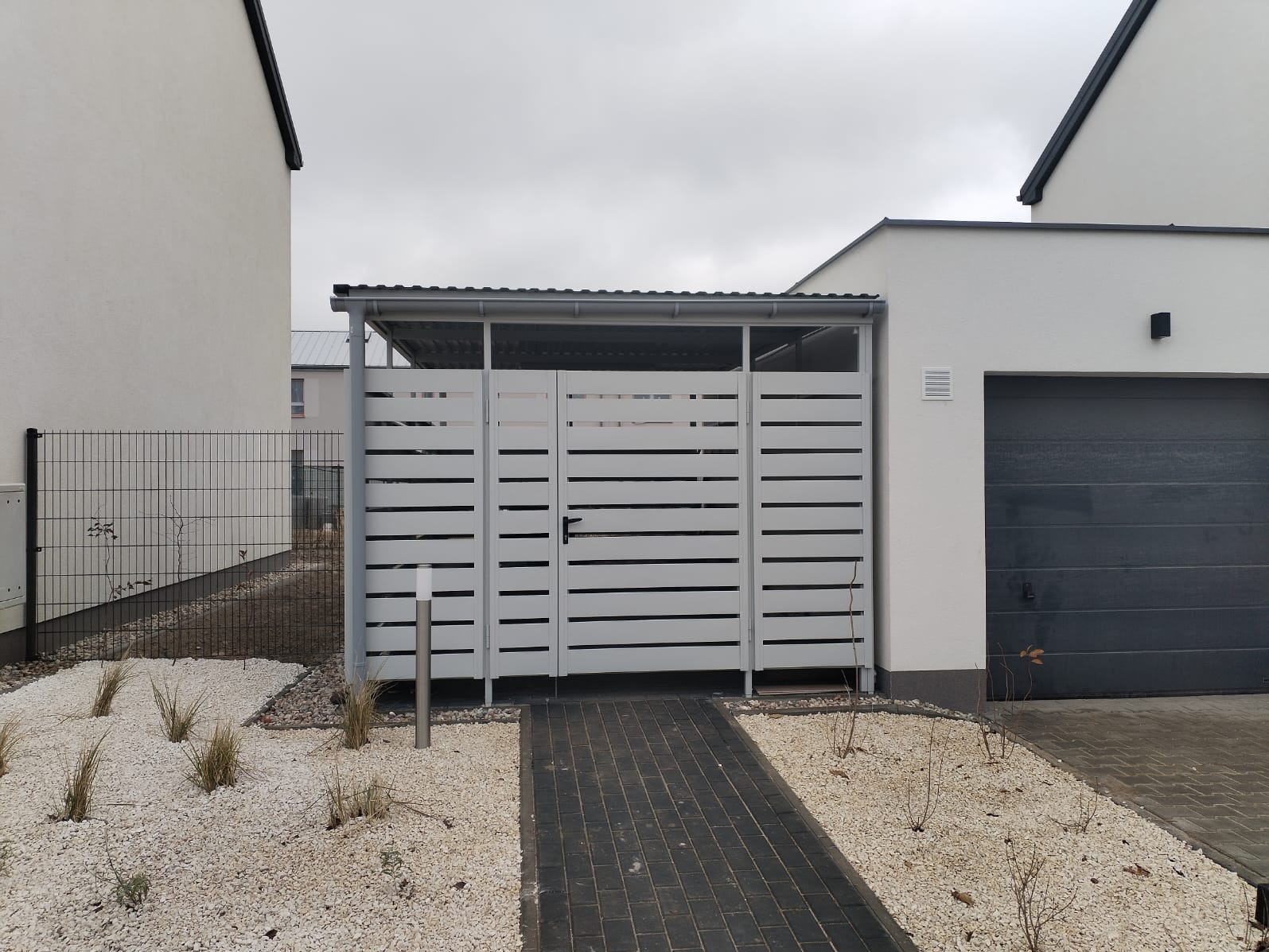 Nowoczesna, szara, ażurowa konstrukcja zadaszenia nad bramą, z poziomymi szczeblami, z czarną klamką, na tle białego domu z szarą bramą garażową, wyłożona kostką i białym żwirem z roślinami.