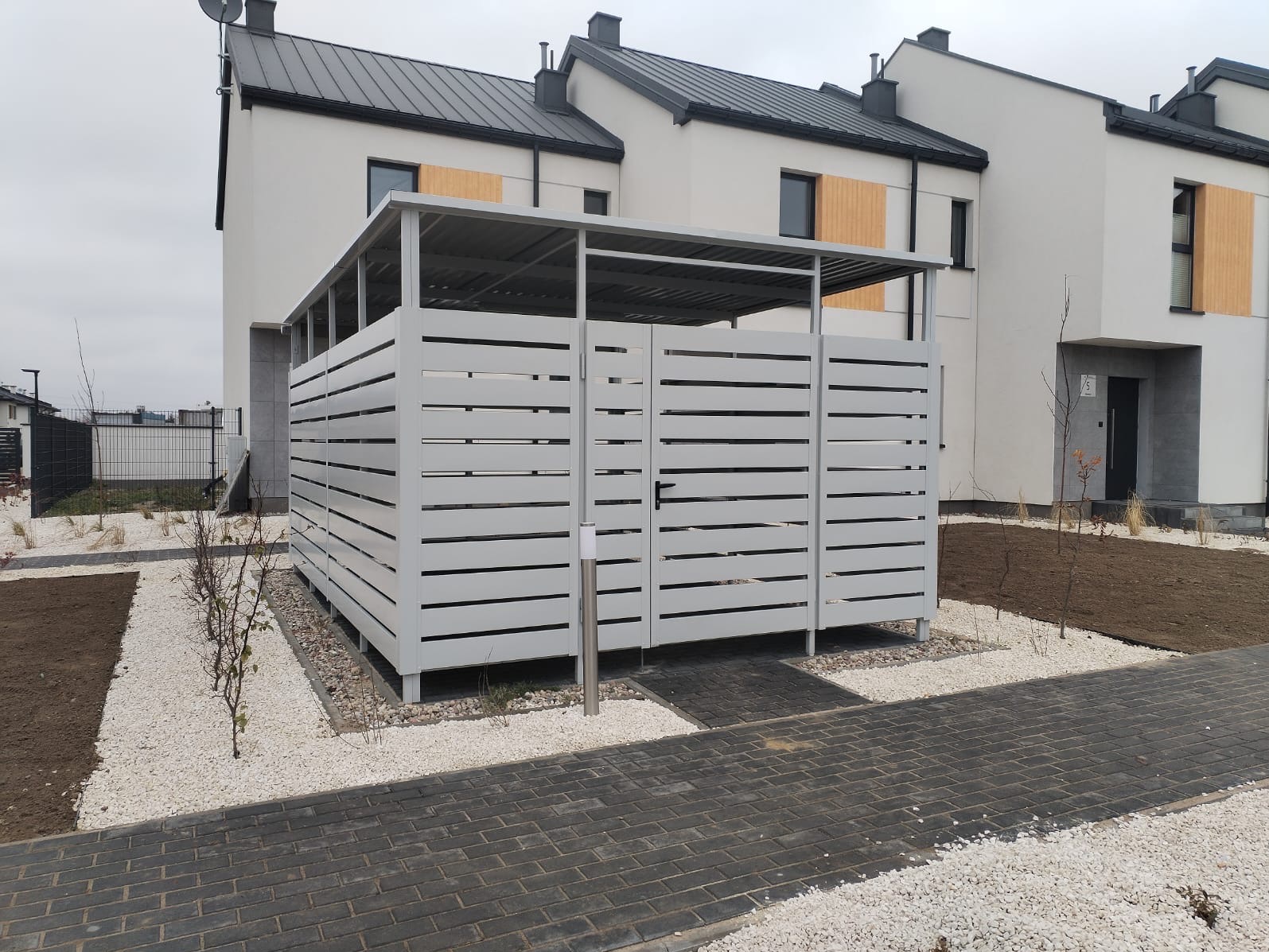 Szara, ażurowa konstrukcja z zadaszeniem na podwórku nowego osiedla, otoczona białym kamieniem i kostką brukową.