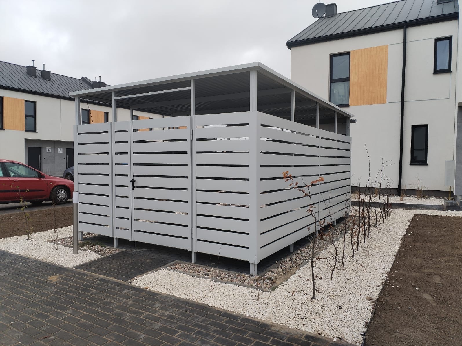 Nowoczesna, biała konstrukcja z poziomych listew z zadaszeniem, prawdopodobnie na śmietnik, na tle domów jednorodzinnych i kostki brukowej.