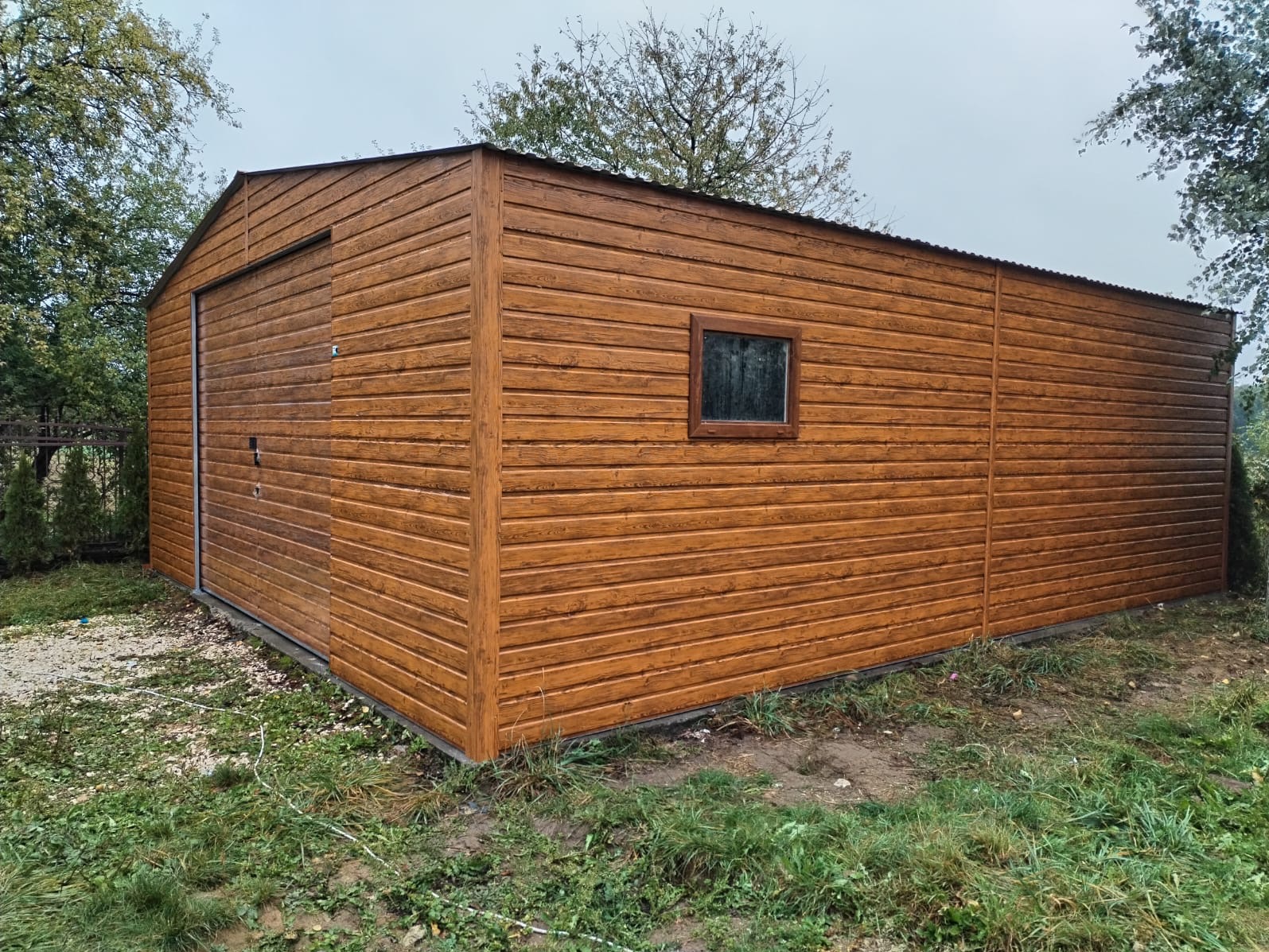 Brązowy garaż z desek z przesuwną bramą i oknem, na tle drzew i trawy.