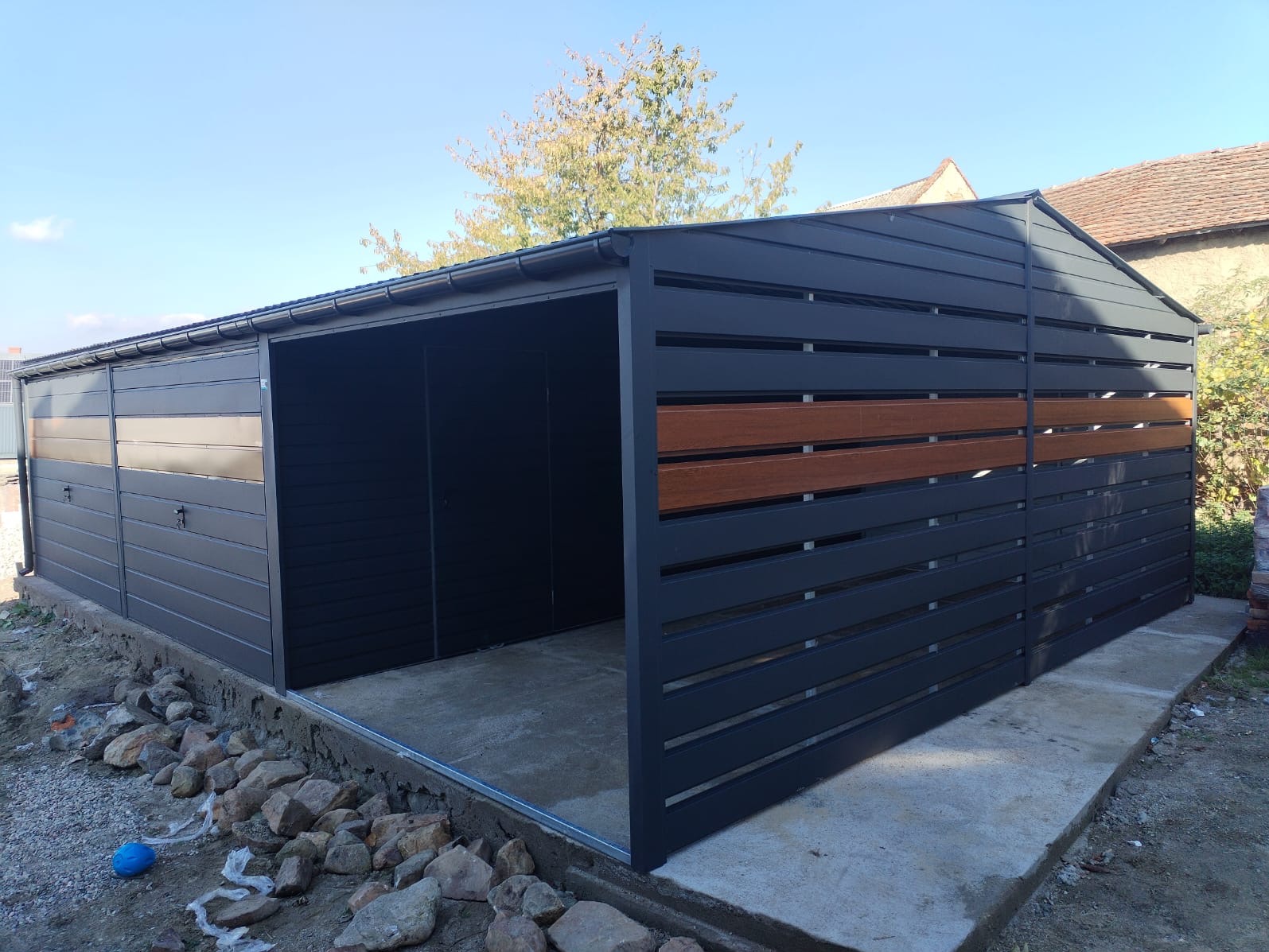 Nowoczesny, antracytowy garaż z elementami dekoracyjnymi z drewna, widoczne kamienie wokół fundamentów, błękitne niebo w tle.