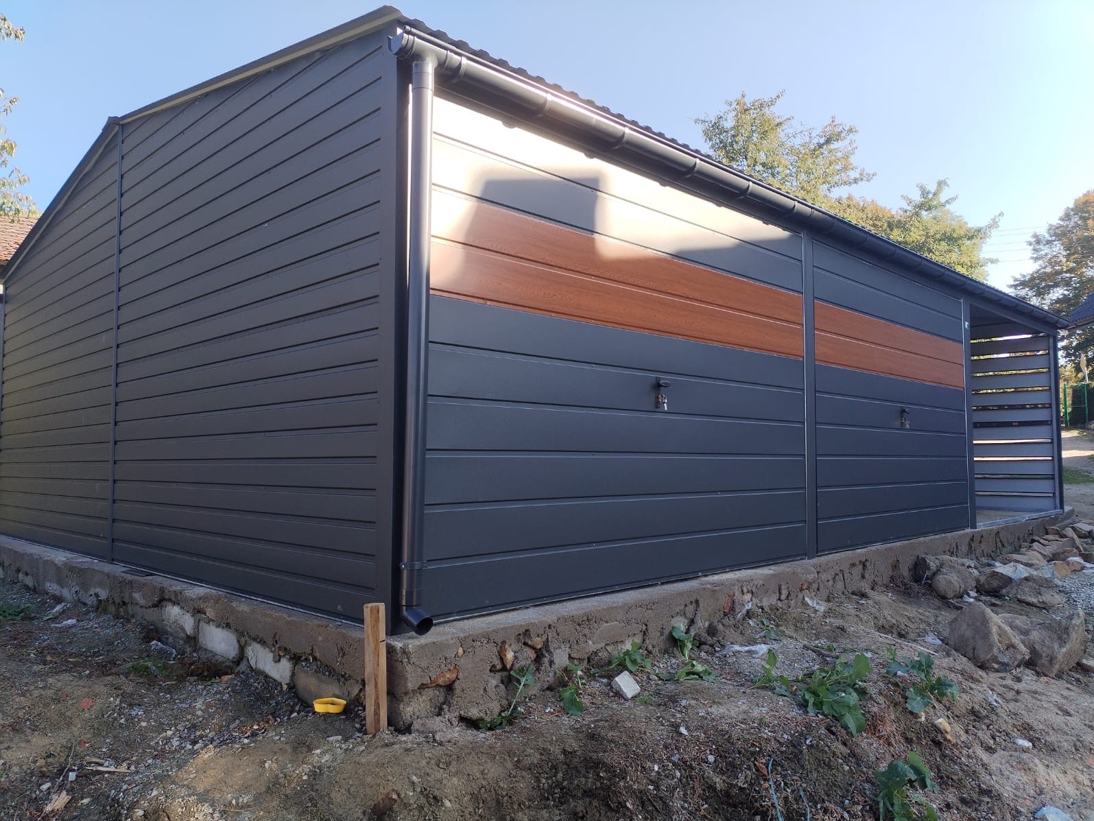 Szary garaż blaszany z poziomymi panelami i brązowym akcentem na górze, widoczny z zewnątrz, z niedokończonym fundamentem betonowym i ziemią wokół.