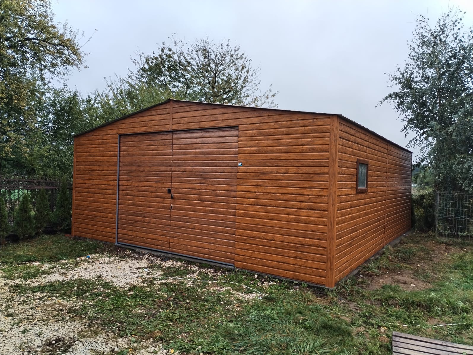 Gotowy, brązowy garaż blaszany z bramą segmentową i małym oknem bocznym, umieszczony na działce z widoczną roślinnością i szarym niebem.