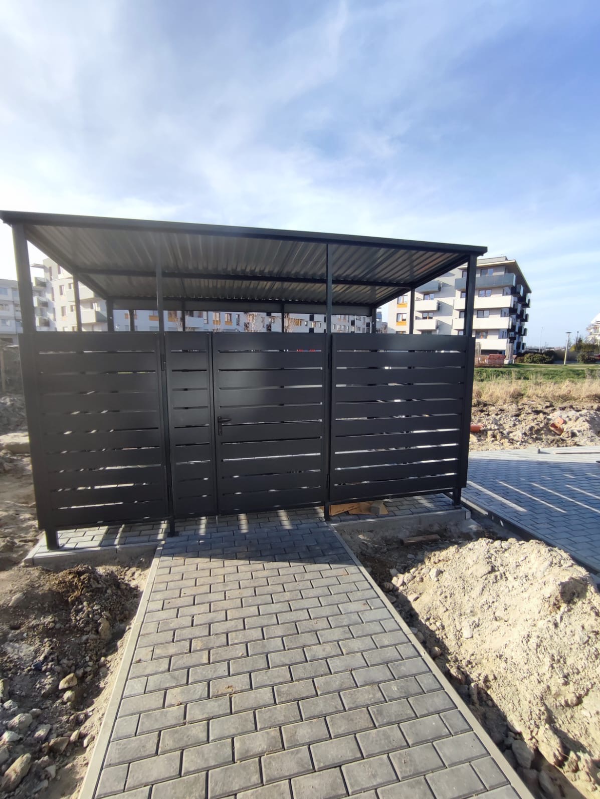 Nowoczesna wiata śmietnikowa z metalową konstrukcją i poziomymi panelami, z kostki brukowej prowadzi do niej chodnik, w tle widoczny budynek mieszkalny.
