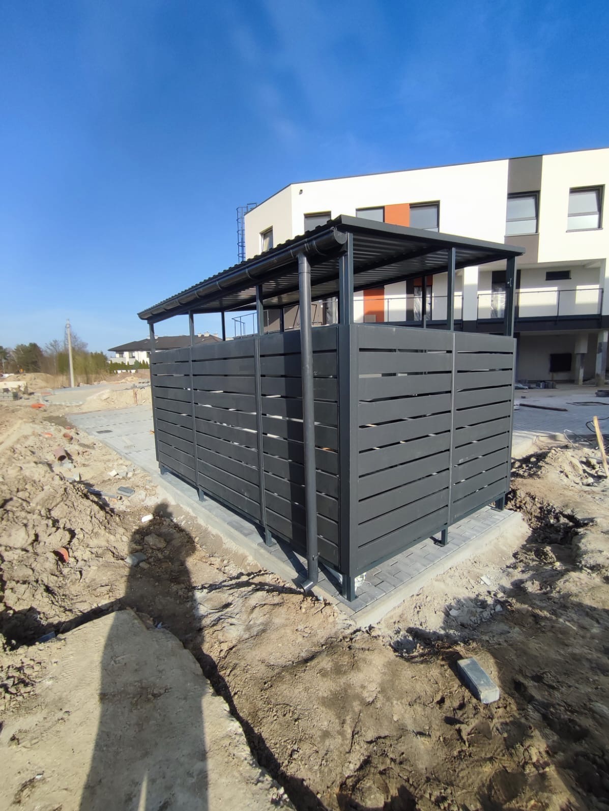 Nowoczesna wiata śmietnikowa z grafitowych paneli, zadaszona, na kostce brukowej, w tle budynek mieszkalny w trakcie wykończenia, ziemia wokół rozkopana.