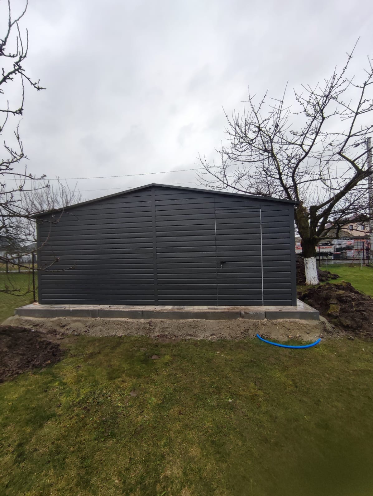 Szary garaż z blachy falistej, z pojedynczymi drzwiami, osadzony na betonowej podmurówce na trawniku, z widocznym niebieskim przewodem i drzewem z bielonym pniem w tle.