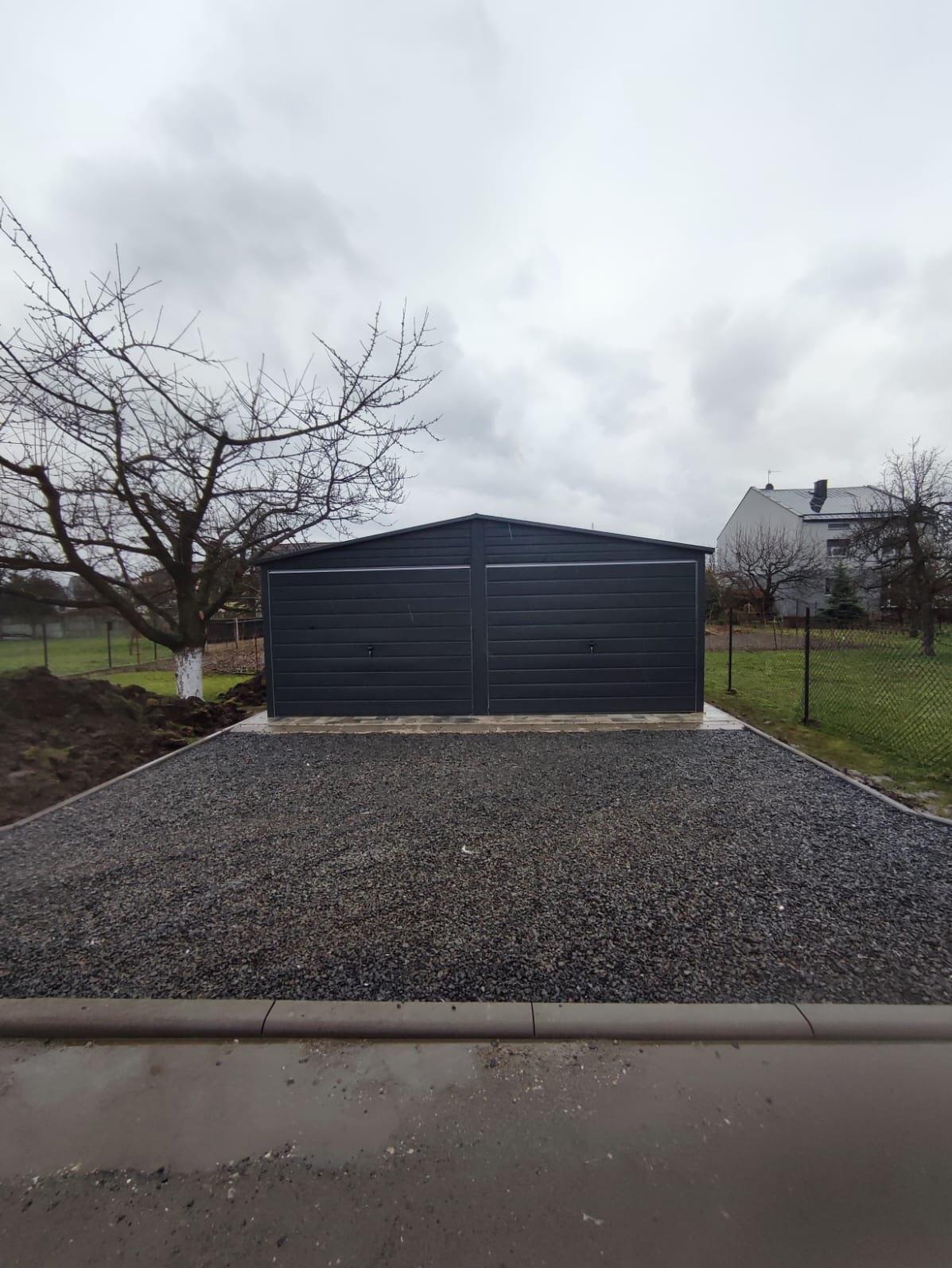 Szary dwustanowiskowy garaż z blachy falistej na betonowej wylewce i podsypce z grysu, widok z zewnątrz w pochmurny dzień.