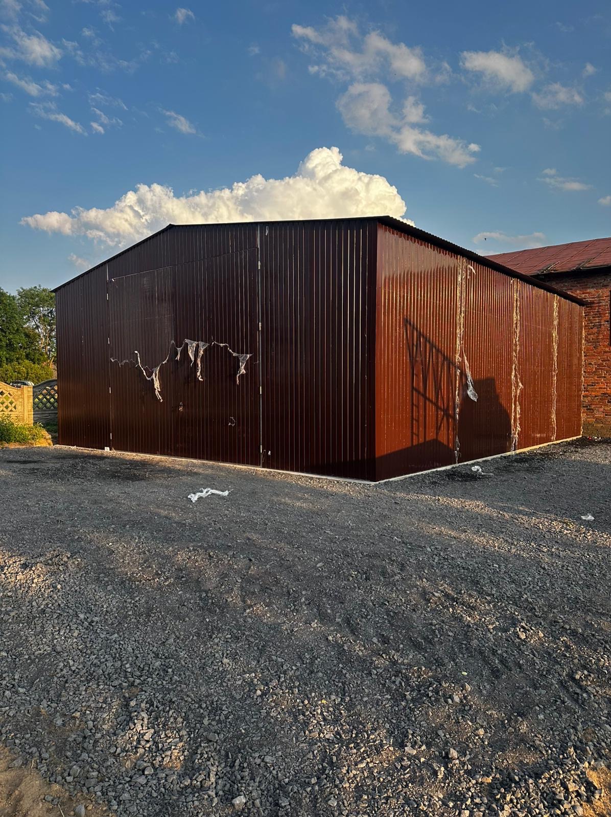 Brązowy garaż z blachy falistej, z resztkami folii ochronnej na ścianach, stojący na żwirowym podłożu, na tle błękitnego nieba z białymi chmurami i fragmentu ceglanego budynku.