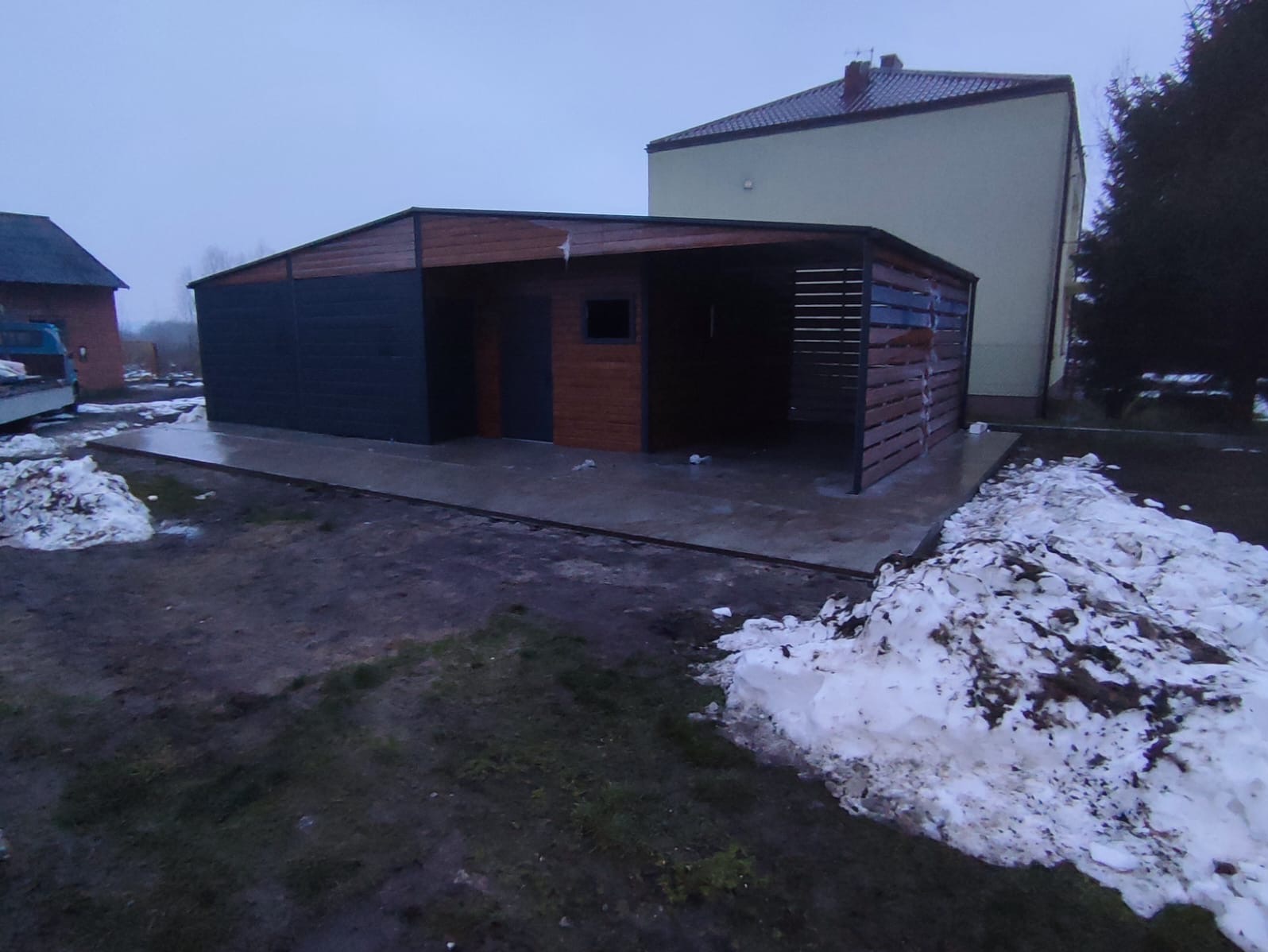 Nowoczesny garaż blaszany z elementami drewnopodobnymi i zadaszoną częścią, widoczny w pochmurny dzień z resztkami śniegu na ziemi.