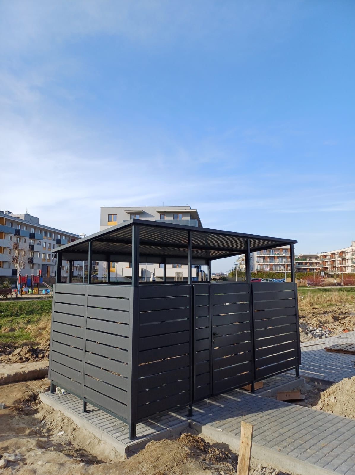 Nowoczesna, grafitowa konstrukcja z poziomych paneli, prawdopodobnie osłona na pojemniki na śmieci, umieszczona na nowo wybrukowanym chodniku w otoczeniu budynków mieszkalnych i placu budowy.