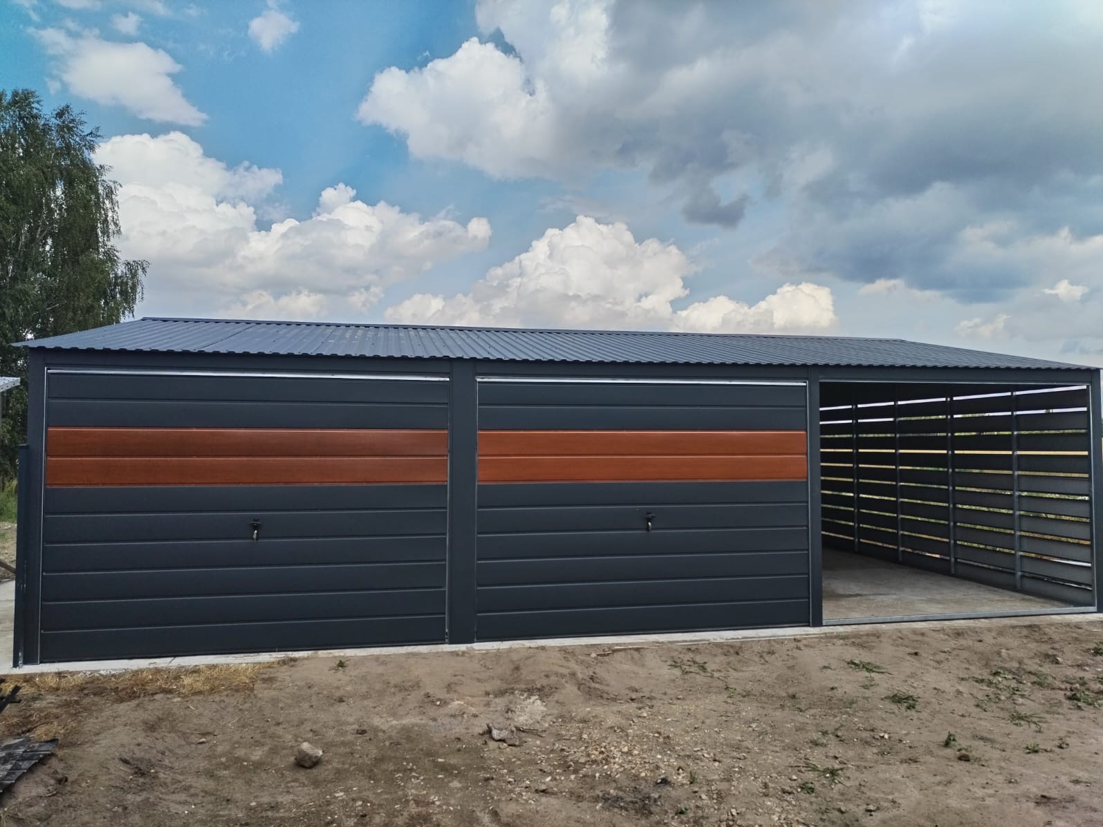 Szary garaż z dwoma segmentowymi bramami z poziomymi panelami, z których jeden jest w kolorze drewna, oraz otwartą wiatą z poziomymi szczeblami, na tle błękitnego nieba z chmurami.
