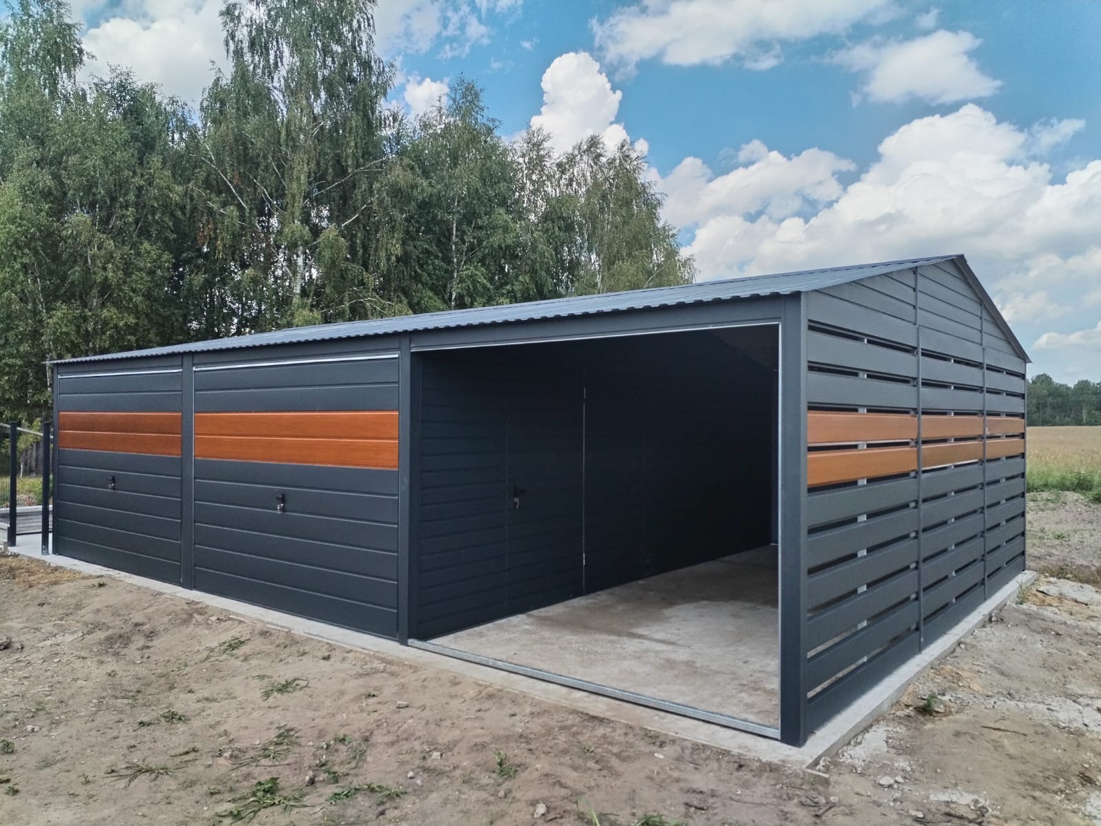 Nowoczesny, grafitowy garaż z elementami dekoracyjnymi imitującymi drewno, z otwartą bramą wjazdową i widocznym betonowym fundamentem, na tle zieleni i błękitnego nieba.
