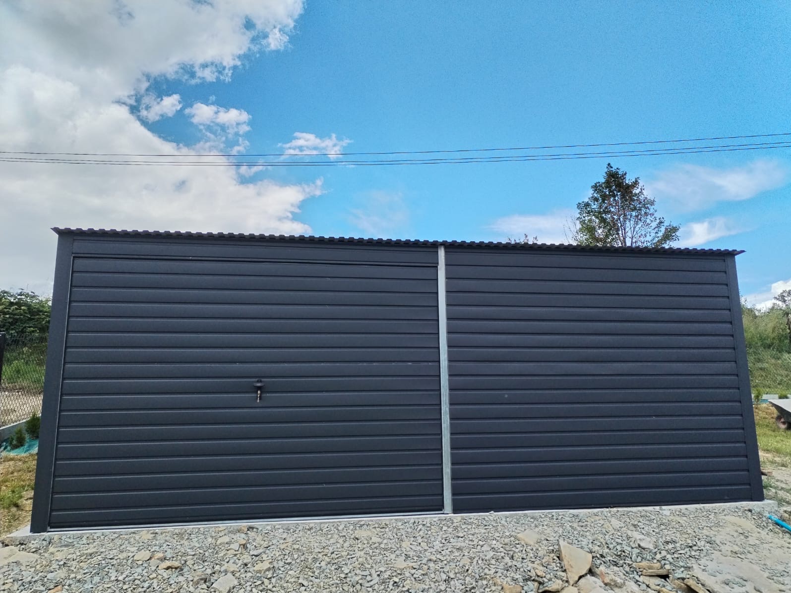 Grafitowy garaż z blachy falistej, widoczny z przodu, zamontowany na żwirowym podłożu pod błękitnym niebem z chmurami.