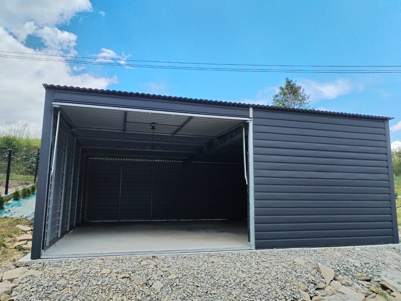 Nowoczesny, grafitowy garaż z blachy falistej z otwartą przesuwną bramą, na żwirowym podłożu, na tle błękitnego nieba z chmurami.