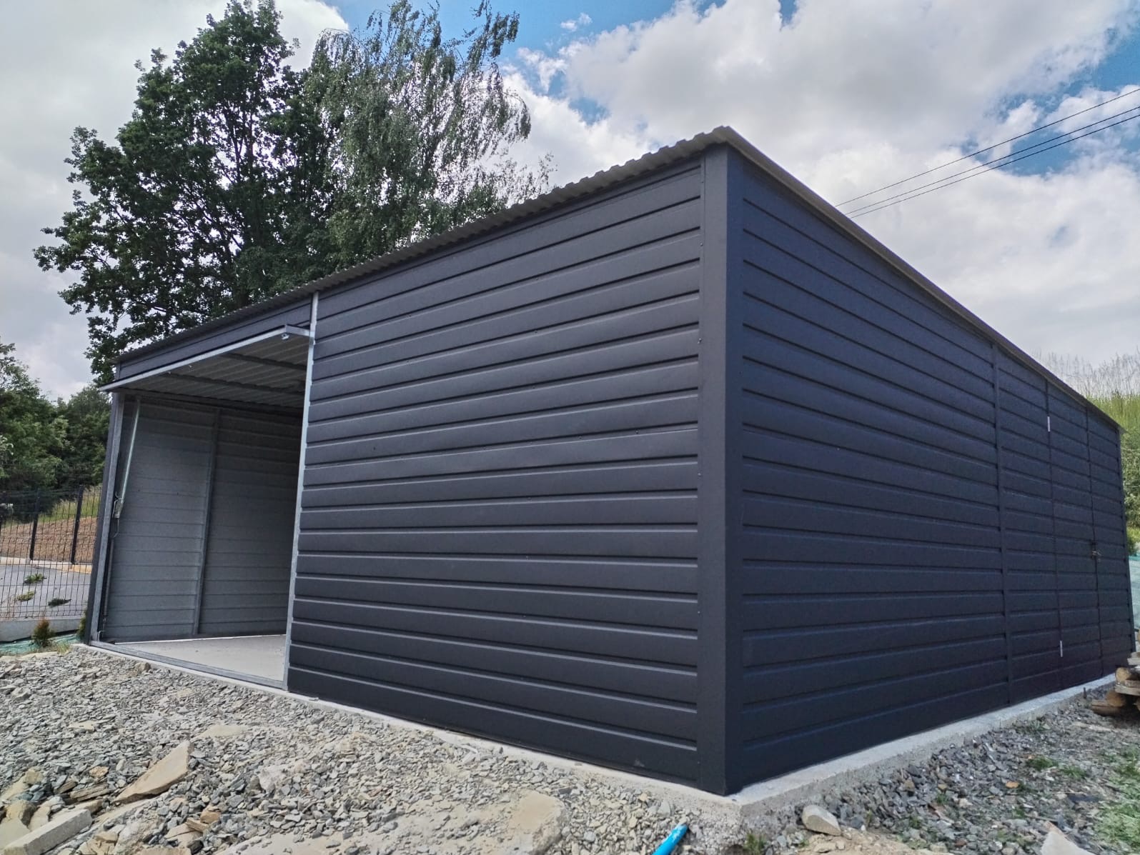 Nowoczesny, ciemnoszary garaż blaszany z wiatą w Stróży, z widoczną konstrukcją stalową i podłożem z kamieni.