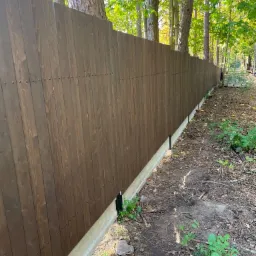 Długi, ciemnobrązowy drewniany płot z pionowych desek, osadzony na betonowej podmur&oacute;wce, biegnący wzdłuż zalesionego terenu.