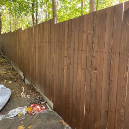 Presto House Olchowik Sp.J. - Długi, brązowy płot z desek z widocznymi śrubami, oddzielający posesję od lasu, z workiem i śmieciami u podstawy.