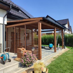 Presto House Olchowik Sp.J. - Drewniana konstrukcja z lamelami, prawdopodobnie w trakcie montażu, widoczne narzędzia i materiały budowlane, na tle domu z panelami słonecznymi i bujnej zieleni.