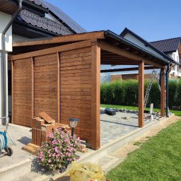 Presto House Olchowik Sp.J. - Drewniana konstrukcja z zadaszeniem, częściowo osłonięta panelami, w trakcie budowy na posesji z trawnikiem i kostką brukową, widoczne narzędzia i materiały budowlane, kwiaty w doniczce.