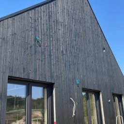 Presto House Olchowik Sp.J. - Częściowy widok zewnętrznej ściany domu z ciemnego drewna z zamontowanymi oknami i widocznymi elementami instalacji elektrycznej. W oknach odbija się wnętrze z materiałami budowlanymi i narzędziami.