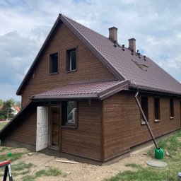 Presto House Olchowik Sp.J. - Brązowy dom z drewnianą elewacją i dachem z blachodachówki, widok z zewnątrz, częściowo otwarte drzwi wejściowe, teren wokół domu w trakcie prac wykończeniowych.