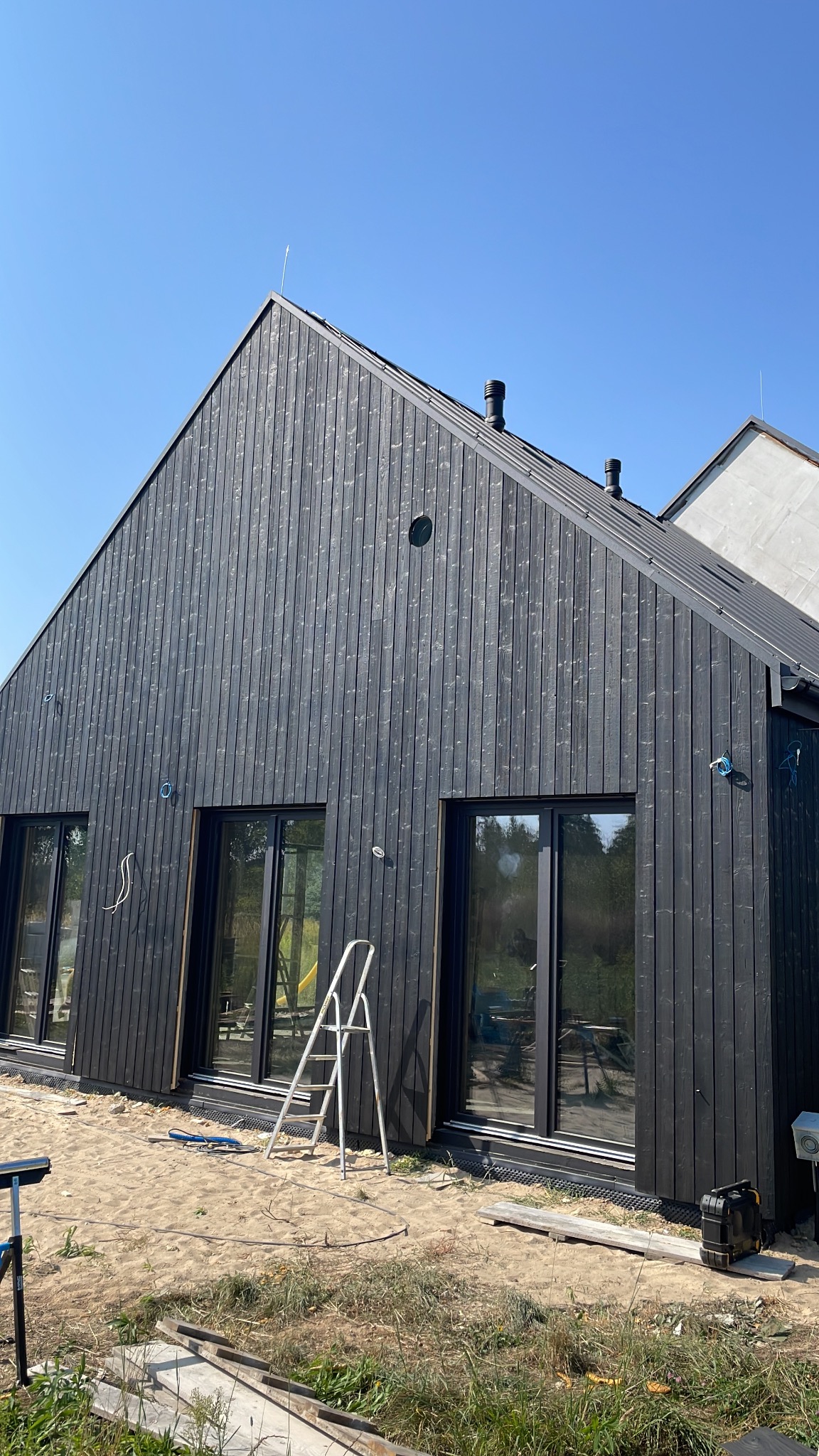 Nowoczesny dom szkieletowy z czarną, drewnianą elewacją w trakcie budowy, widoczne okna, drabina oparta o ścianę i piasek wokół budynku.