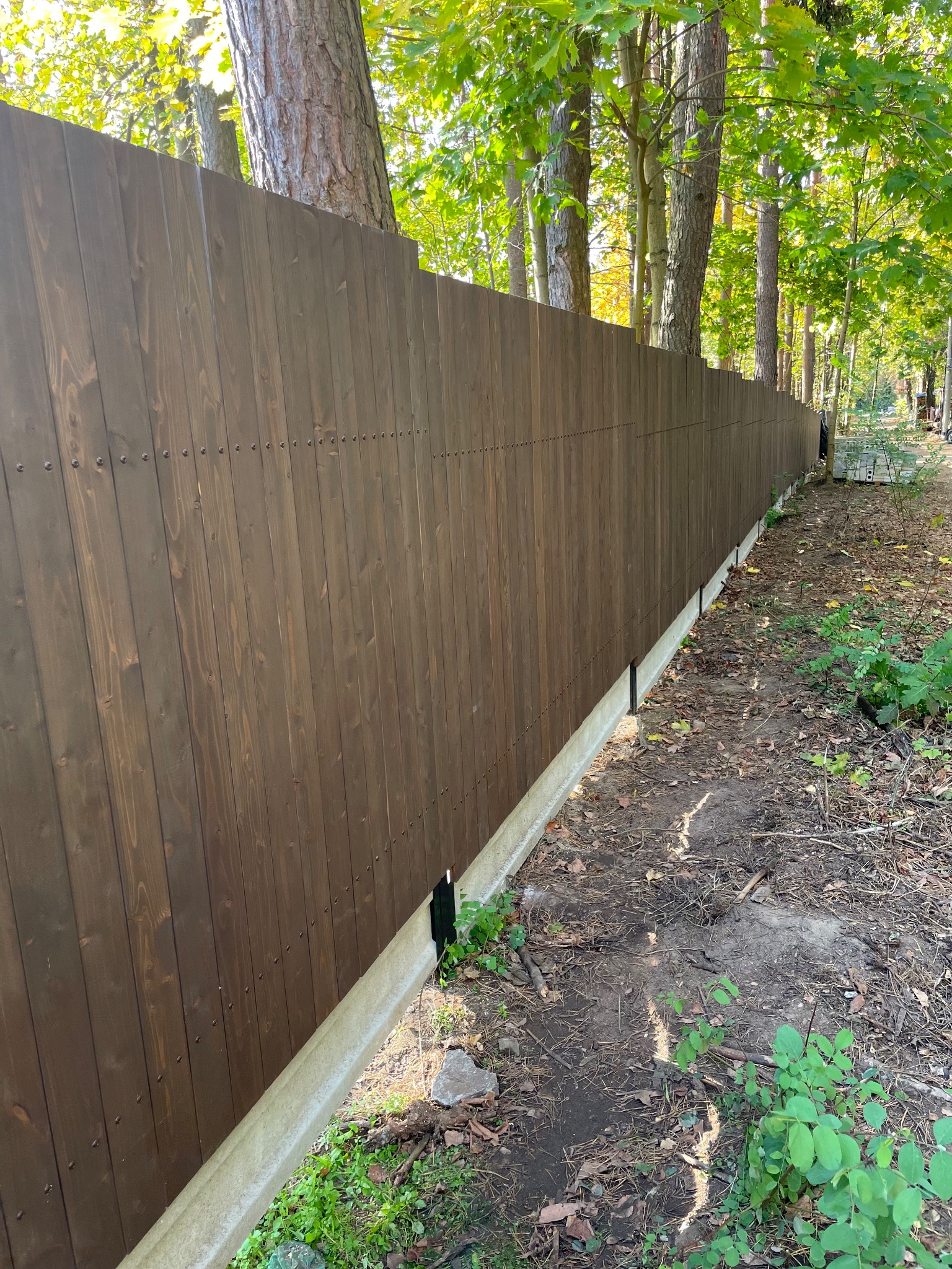 Długi, ciemnobrązowy drewniany płot z pionowych desek, osadzony na betonowej podmurówce, biegnący wzdłuż zalesionego terenu.