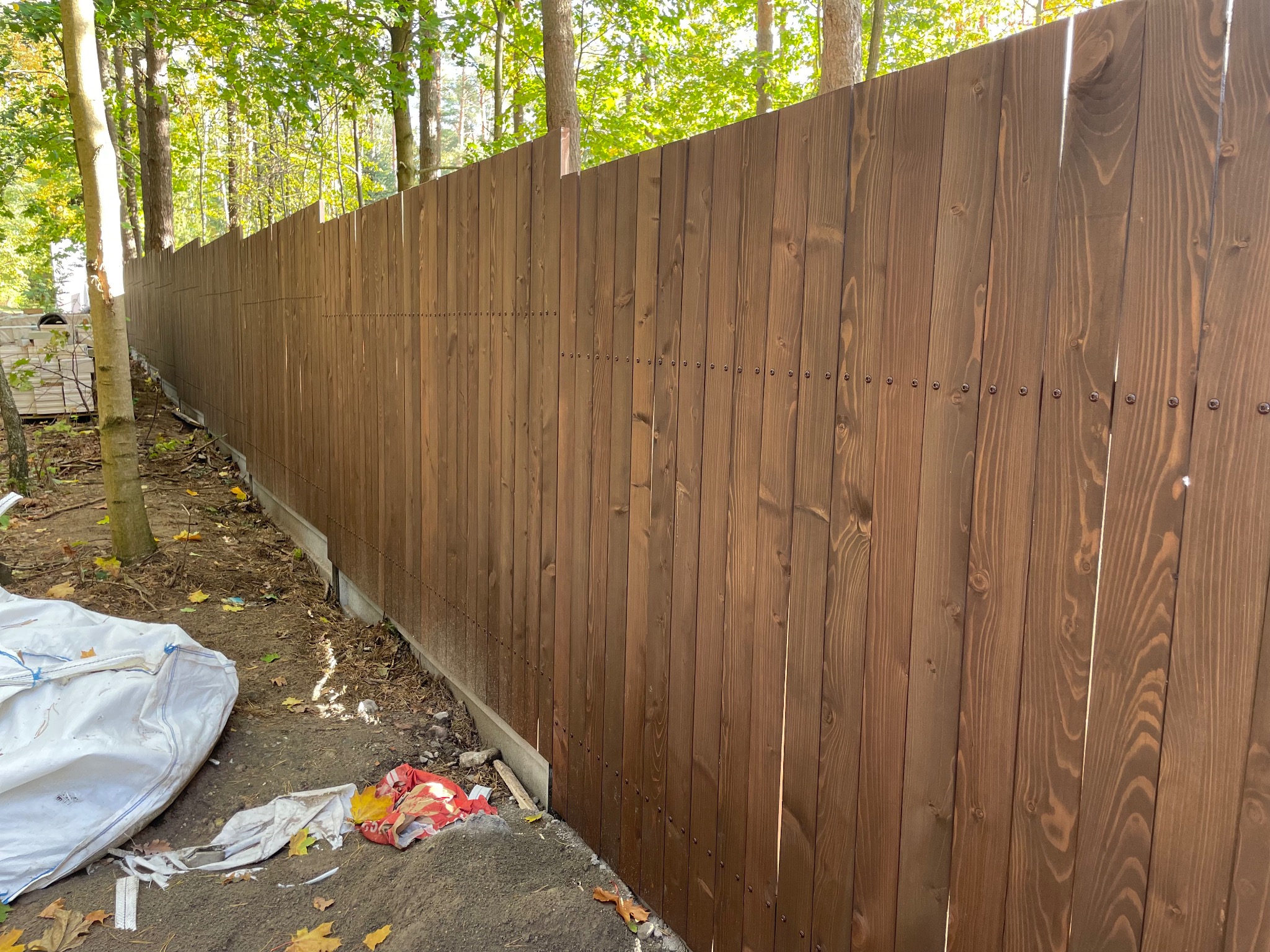 Długi, brązowy płot z desek z widocznymi śrubami, oddzielający posesję od lasu, z workiem i śmieciami u podstawy.