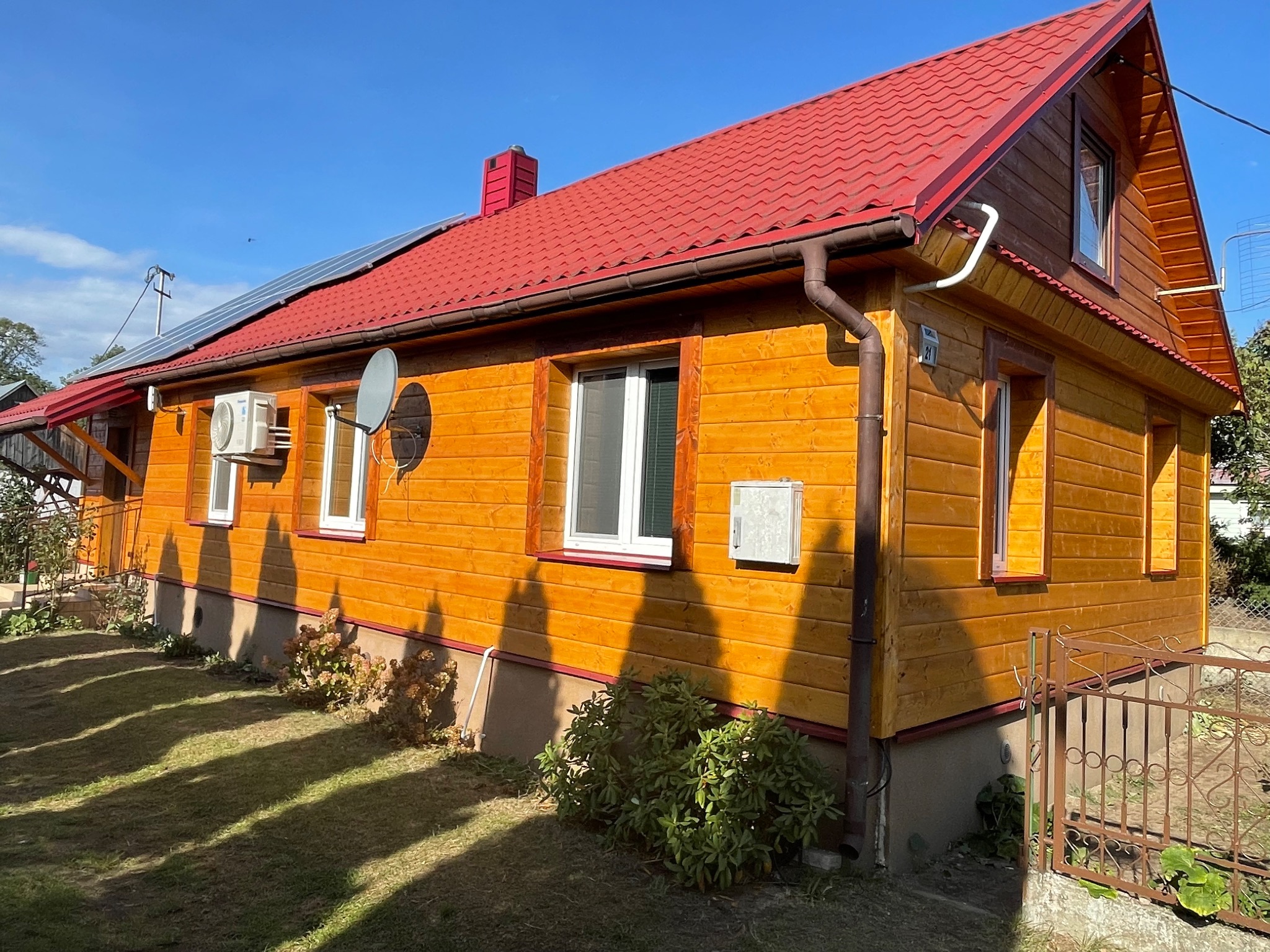 Żółty dom z drewna z czerwoną dachówką, panelami słonecznymi, klimatyzatorem i anteną satelitarną w słoneczny dzień.