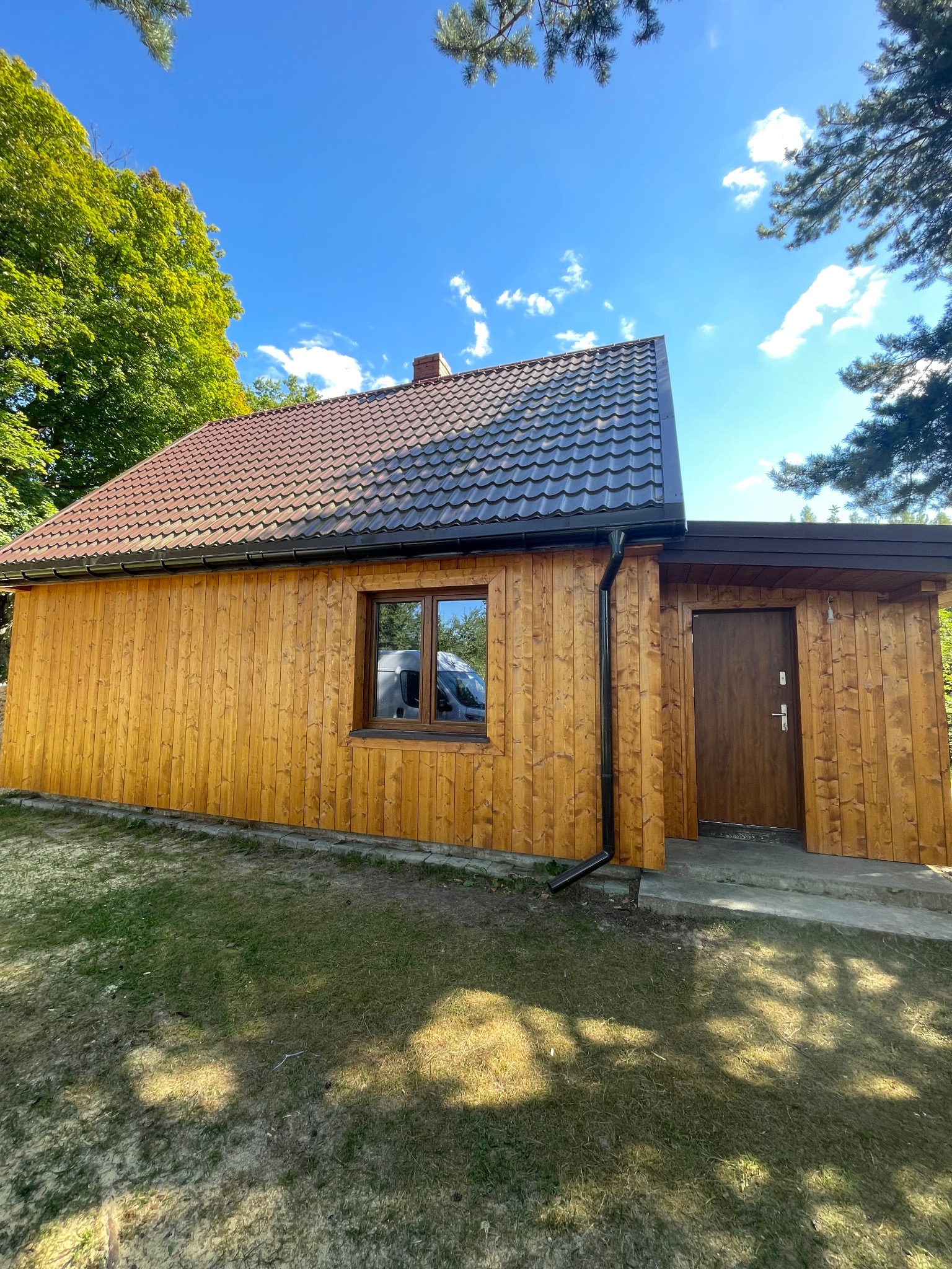 Gotowy budynek z drewnianą elewacją w pionowe deski, brązowy dach z rynną, okno z odbiciem samochodu, brązowe drzwi wejściowe, trawa i drzewa w tle, słoneczny dzień.