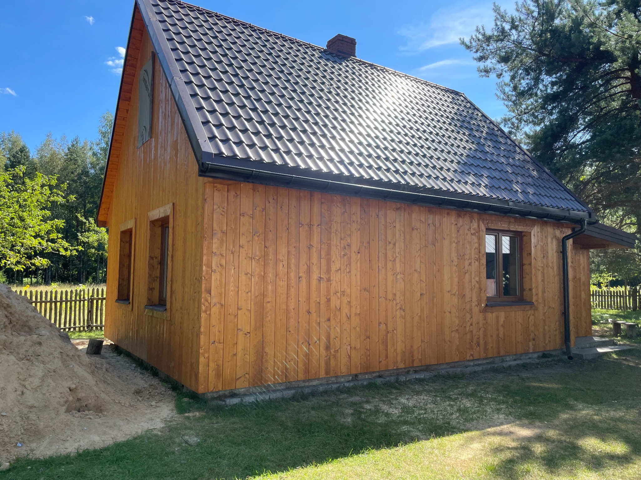 Nowoczesny dom z drewnianą elewacją, ciemnym dachem i widocznym kominem, częściowo otoczony zielenią, z kopcem piasku obok, sugerującym trwające prace wykończeniowe.