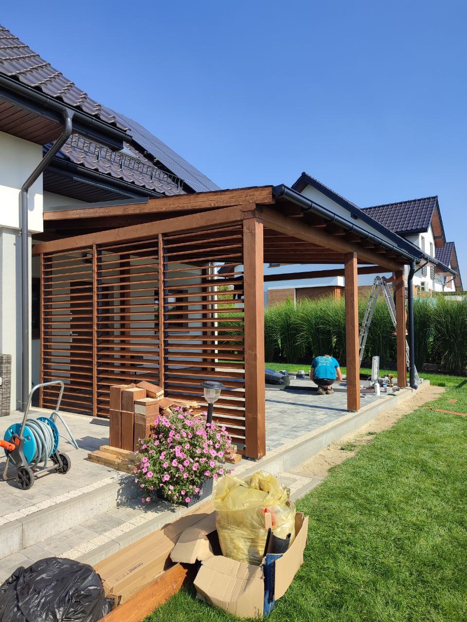 Drewniana konstrukcja z lamelami, prawdopodobnie w trakcie montażu, widoczne narzędzia i materiały budowlane, na tle domu z panelami słonecznymi i bujnej zieleni.