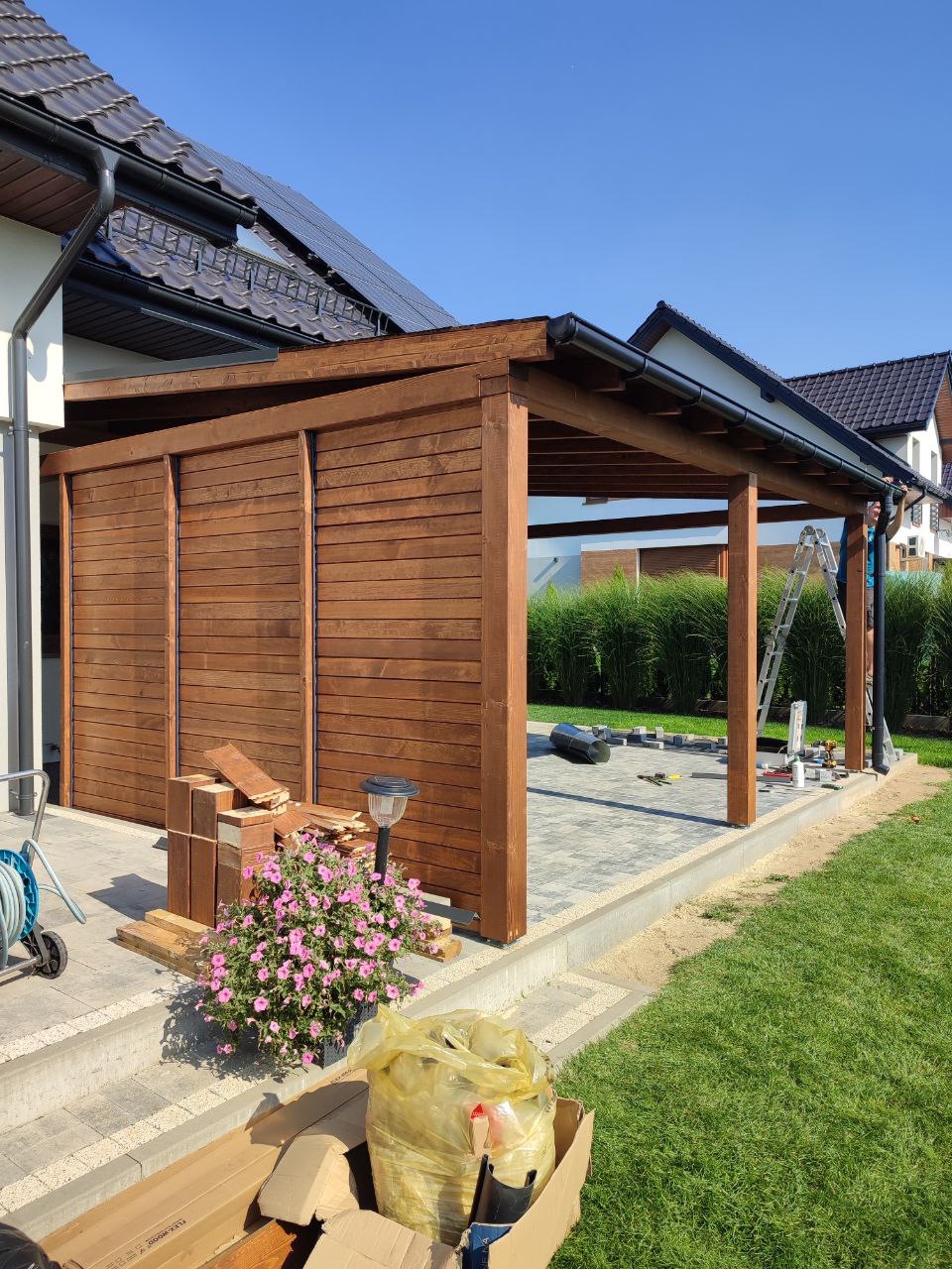 Drewniana konstrukcja z zadaszeniem, częściowo osłonięta panelami, w trakcie budowy na posesji z trawnikiem i kostką brukową, widoczne narzędzia i materiały budowlane, kwiaty w doniczce.