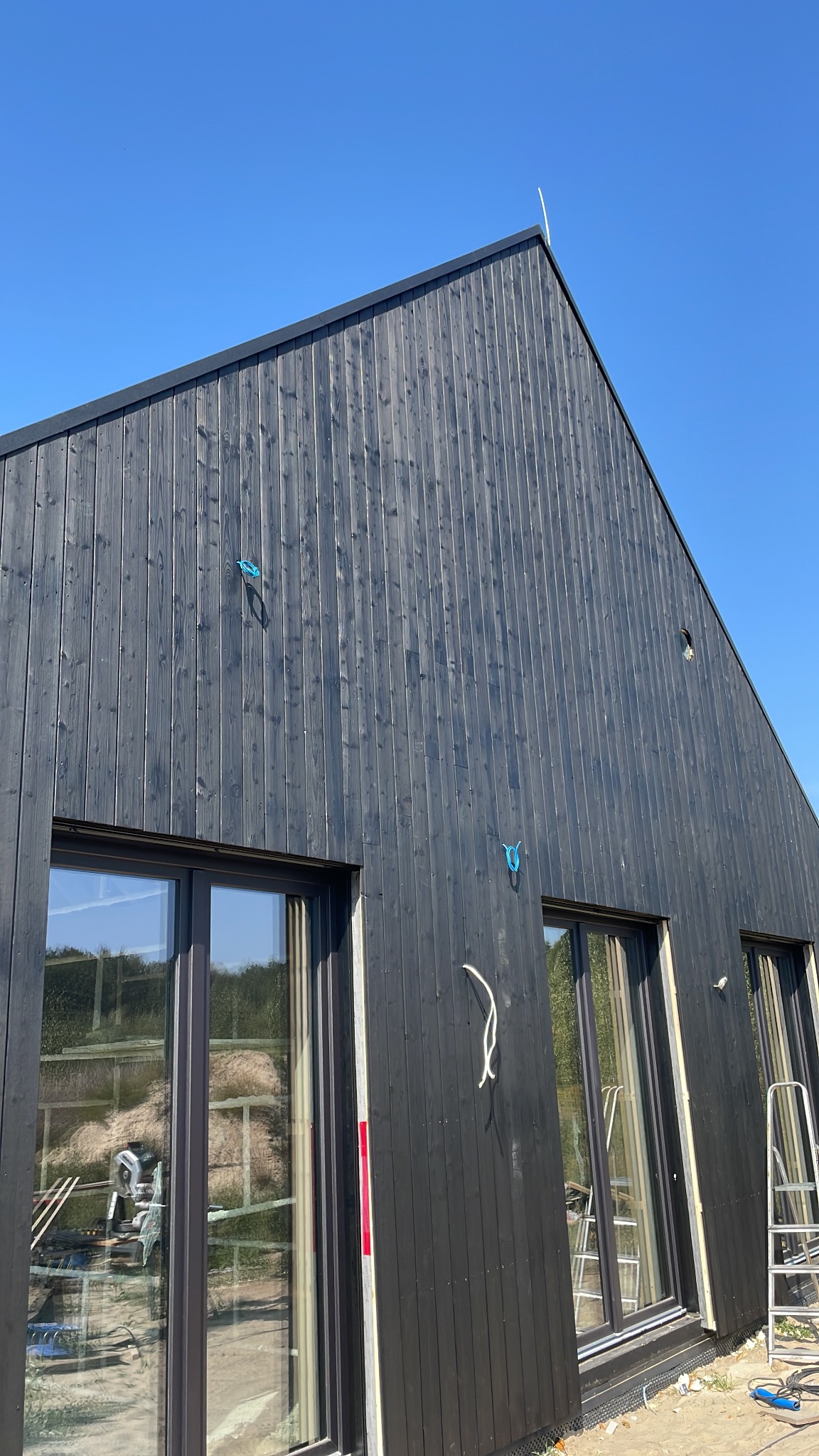 Częściowy widok zewnętrznej ściany domu z ciemnego drewna z zamontowanymi oknami i widocznymi elementami instalacji elektrycznej. W oknach odbija się wnętrze z materiałami budowlanymi i narzędziami.