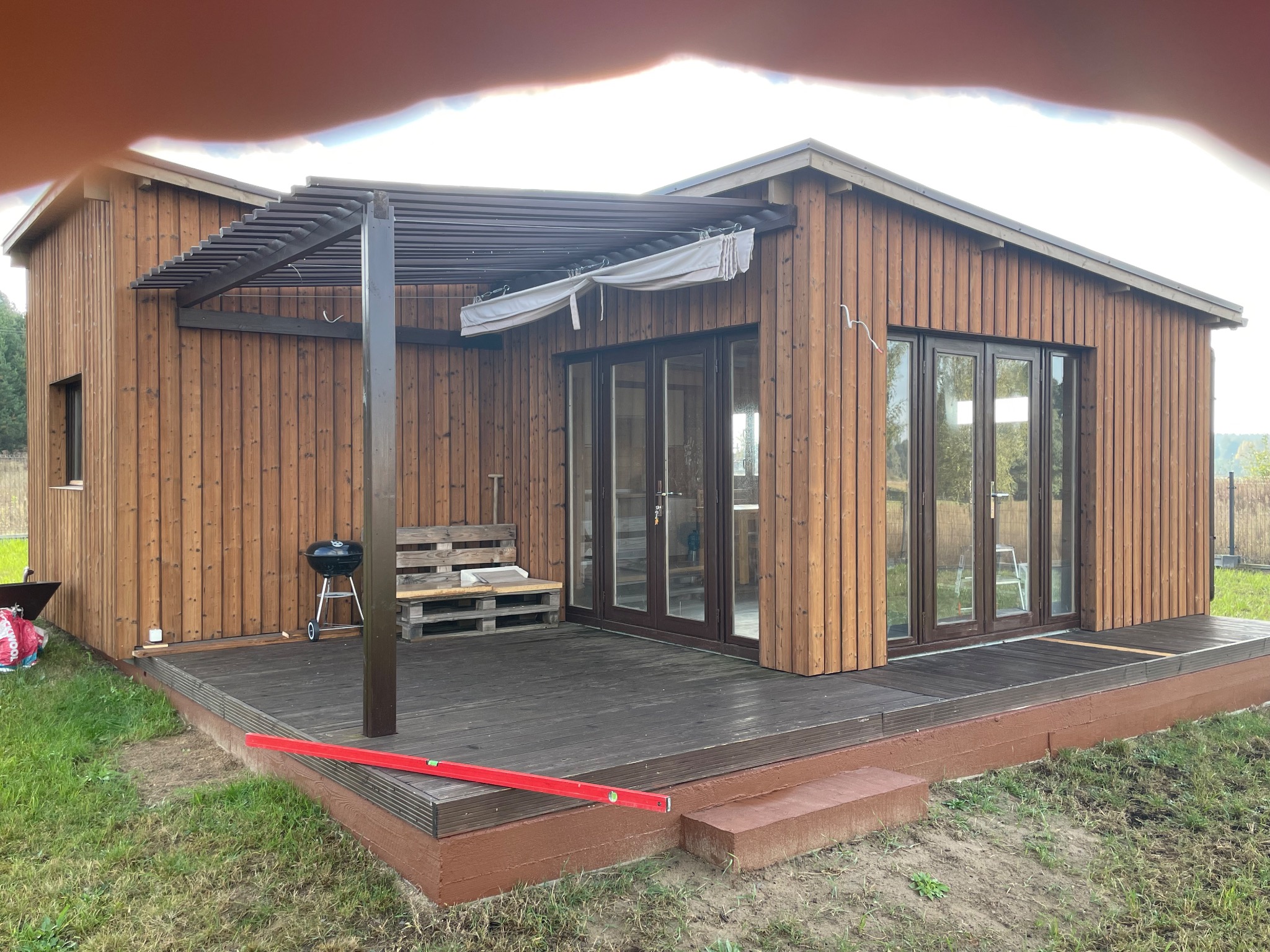 Nowoczesny dom z drewnianą elewacją i tarasem, uzupełniony pergolą z brązowymi elementami, grill i ławka z palet na zewnątrz, poziomica na tarasie.