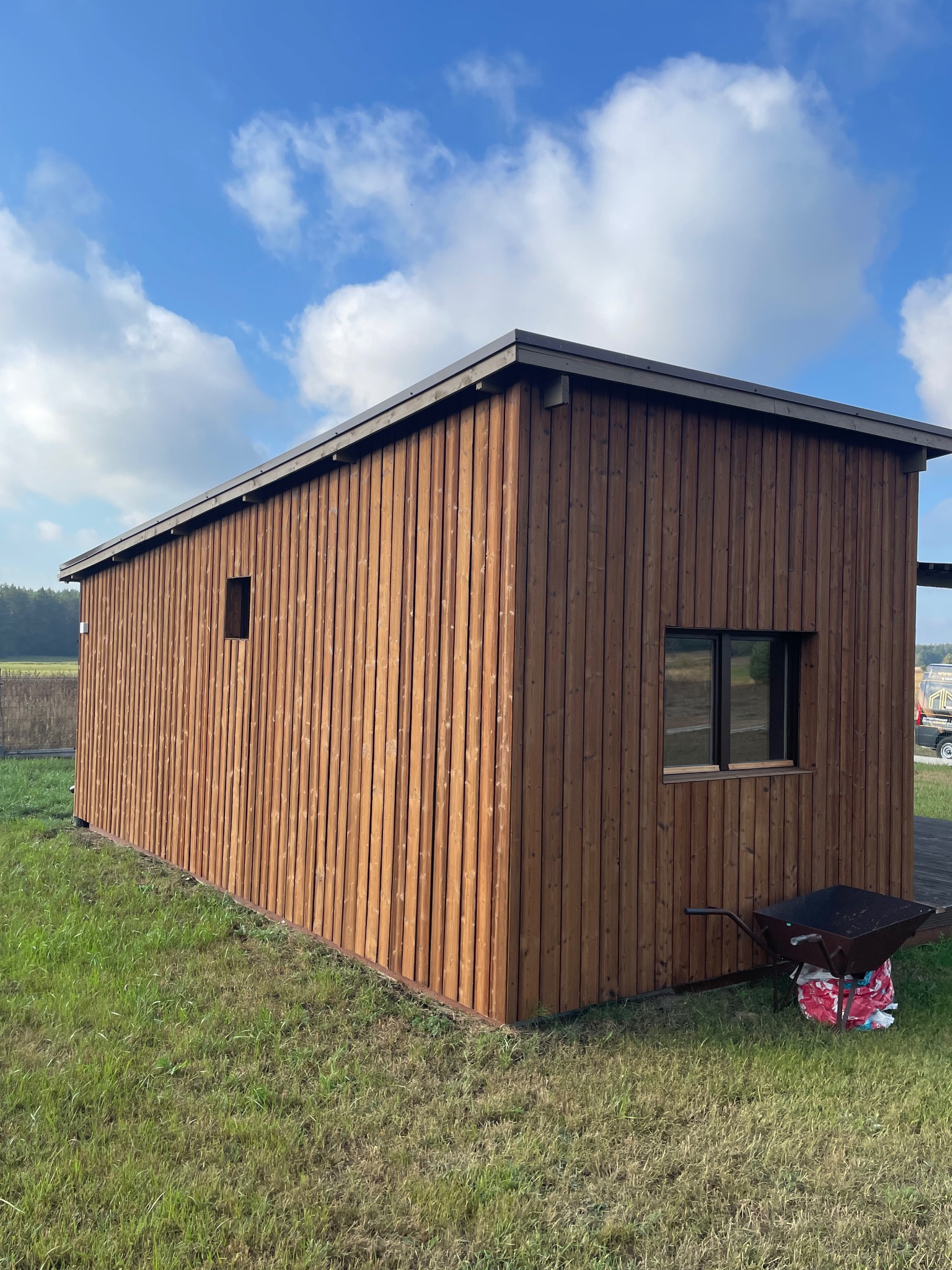 Prosty, parterowy budynek o drewnianej elewacji z pionowych desek, z małymi oknami, na trawiastym terenie, z taczką opartą o ścianę, pod błękitnym niebem z chmurami.