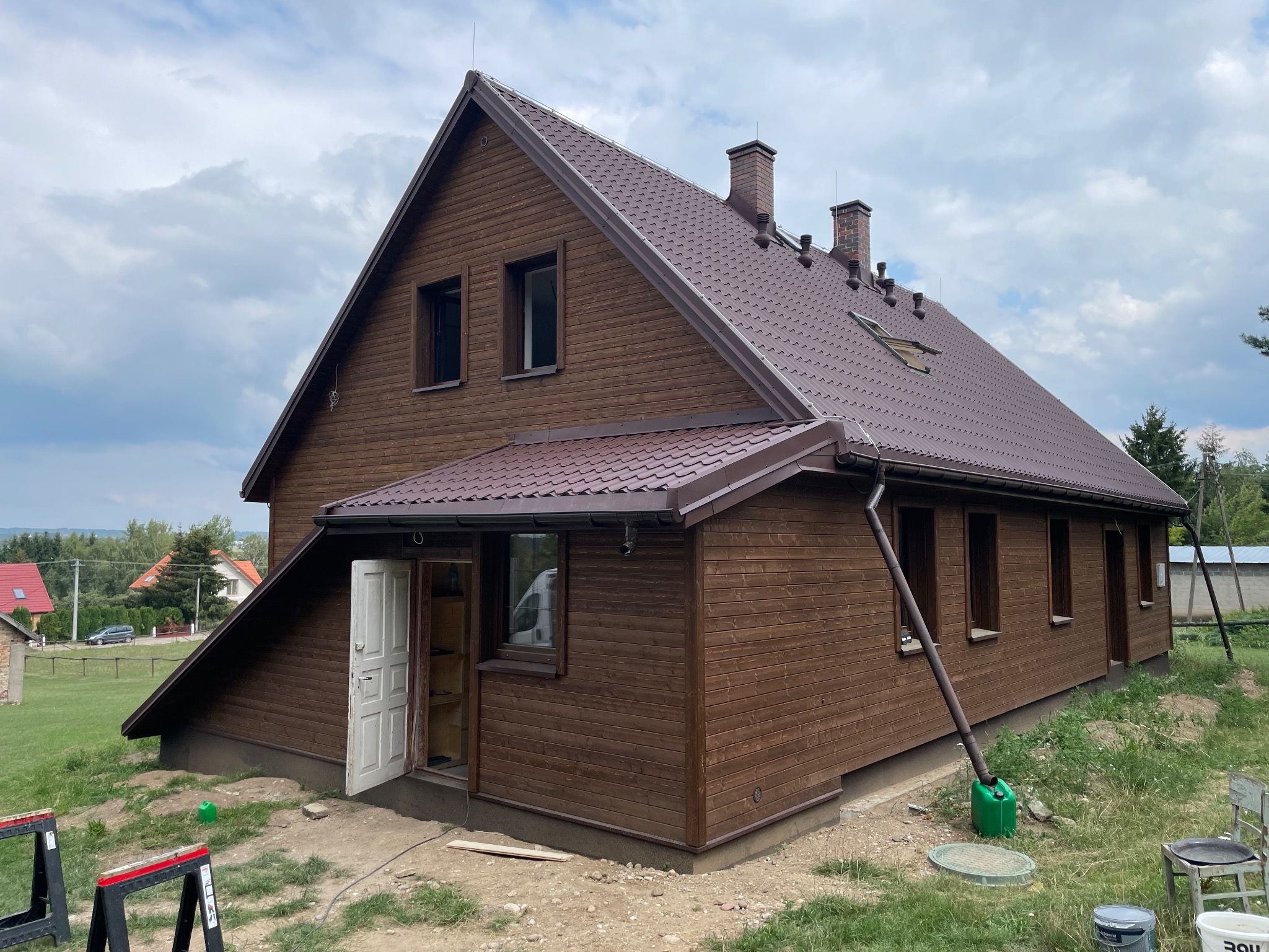 Brązowy dom z drewnianą elewacją i dachem z blachodachówki, widok z zewnątrz, częściowo otwarte drzwi wejściowe, teren wokół domu w trakcie prac wykończeniowych.