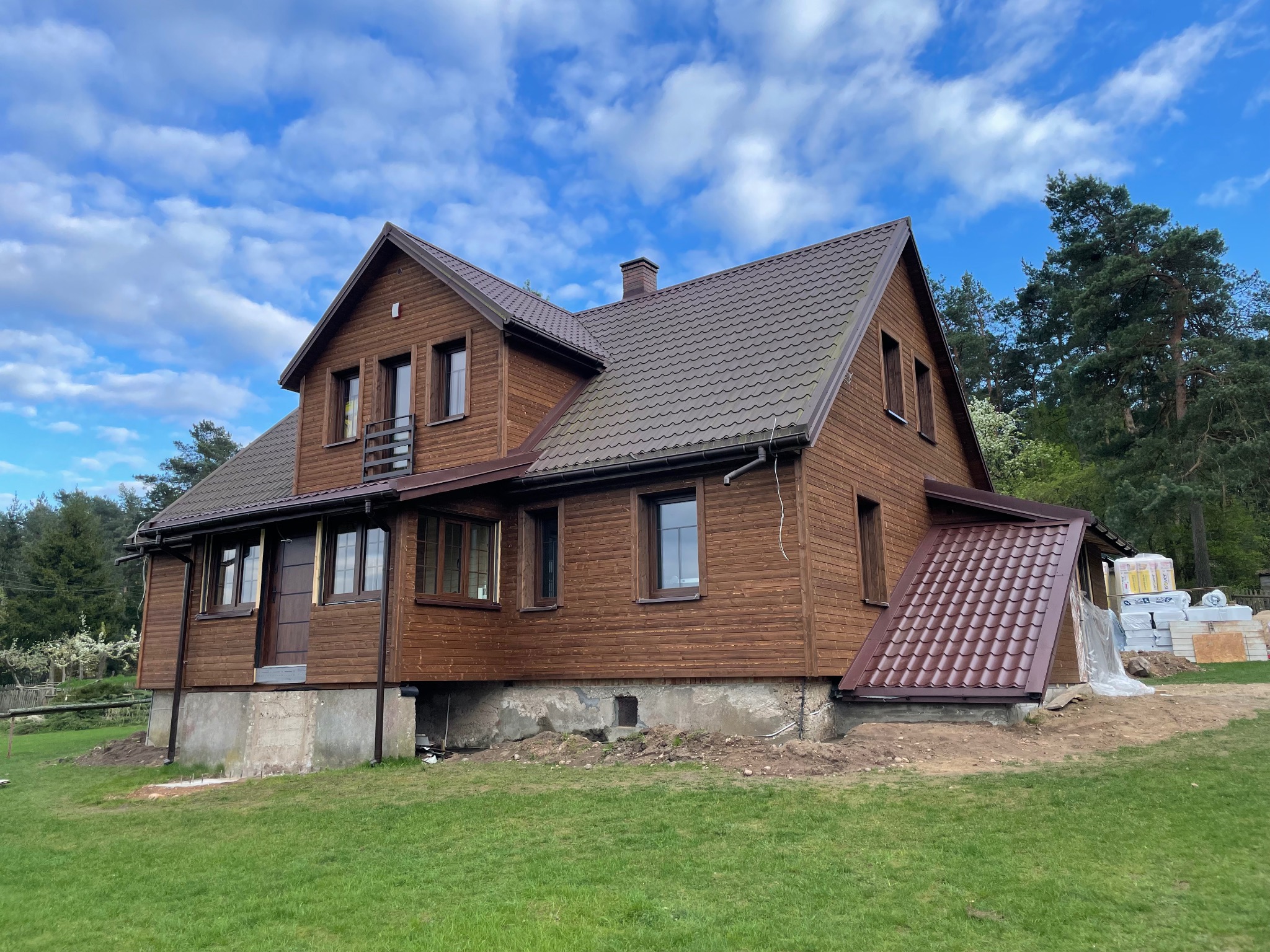 Dwukondygnacyjny dom z brązowymi drewnianymi ścianami i ciemnobrązowym dachem z blachodachówki, częściowo posadowiony na betonowym fundamencie, z widocznymi materiałami budowlanymi obok przybudówki.