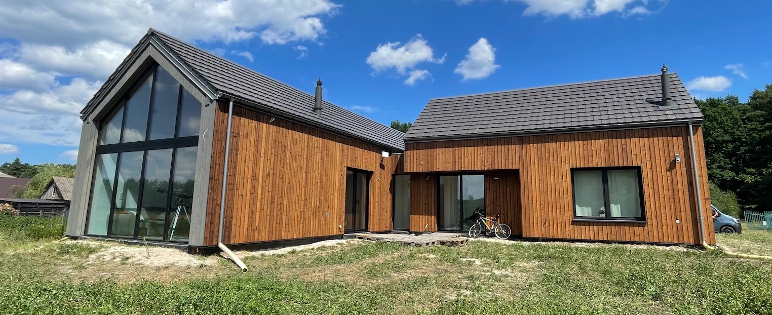 Nowoczesny dom z drewnianą elewacją i dużymi oknami, widoczny rower oparty o ścianę, błękitne niebo w tle.