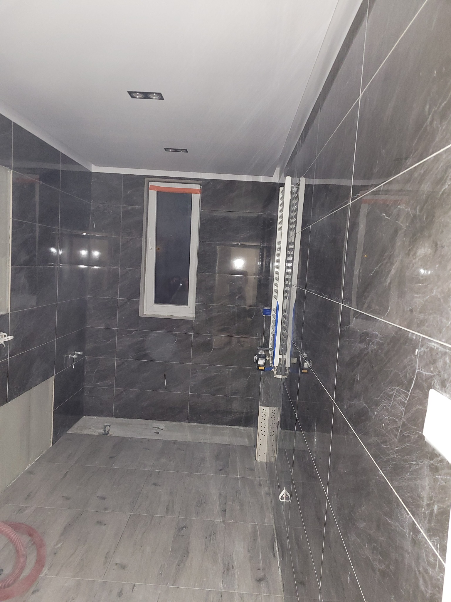 Wnętrze łazienki w trakcie prac wykończeniowych; widoczne szare płytki imitujące marmur na ścianach i podłodze, okno z pomarańczową ramą, oraz instalacja wodno-kanalizacyjna na ścianie.