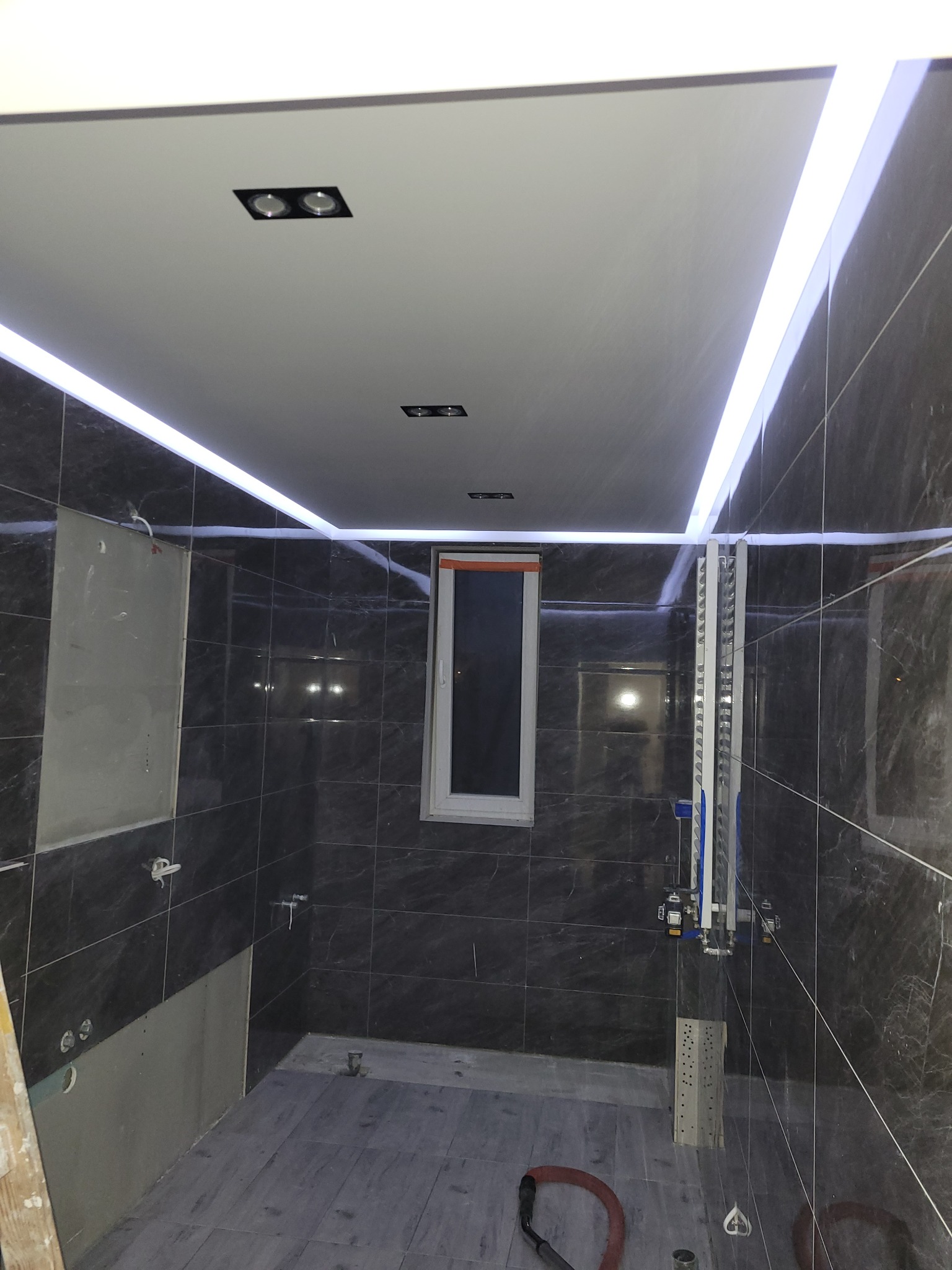 Ciemna łazienka w trakcie remontu, widoczne płytki imitujące marmur na ścianach, podświetlany sufit z punktowymi lampami, okno oraz instalacja wodno-kanalizacyjna w trakcie montażu.