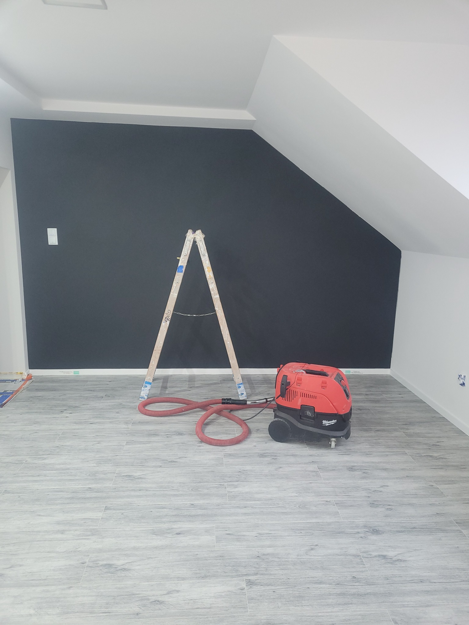 Pokój na poddaszu z pomalowaną na czarno ścianą, drewnianą drabiną i odkurzaczem przemysłowym Milwaukee na podłodze z szarych płytek imitujących drewno.