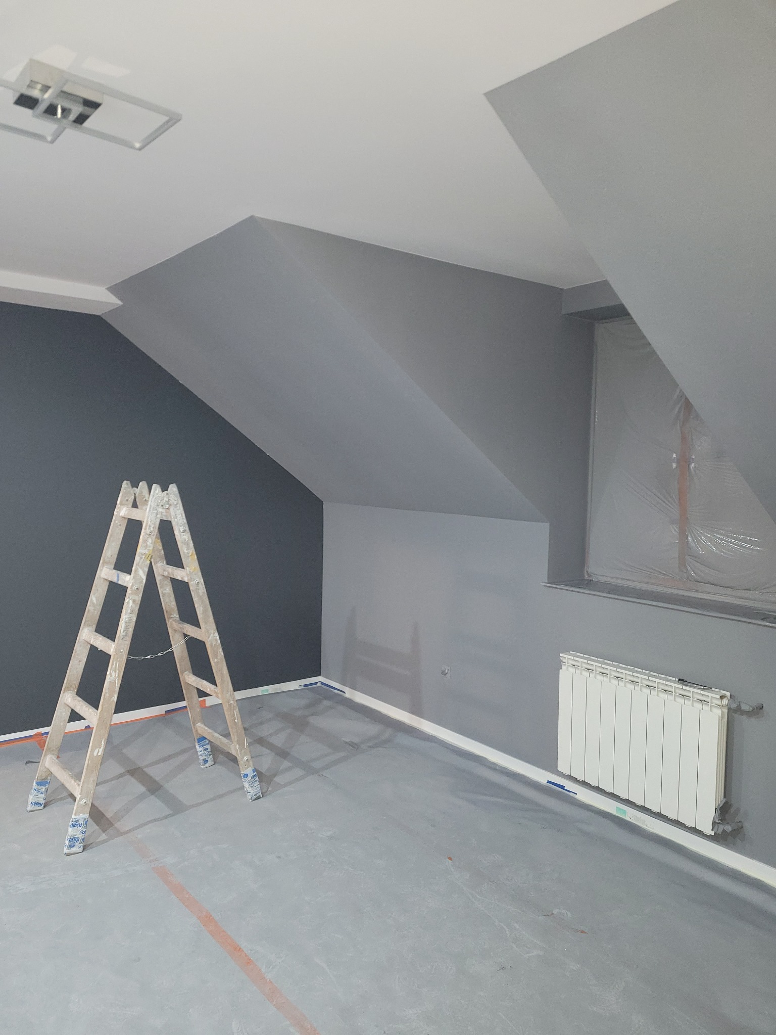 Pokój na poddaszu po malowaniu na szaro, z drewnianą drabiną, zabezpieczonym oknem folią i grzejnikiem.