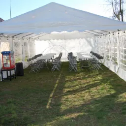 Biały namiot eventowy z oknami, oświetlony girlandami, z ustawionymi stołami i krzesłami, oraz maszyną do popcornu, na trawiastym terenie.