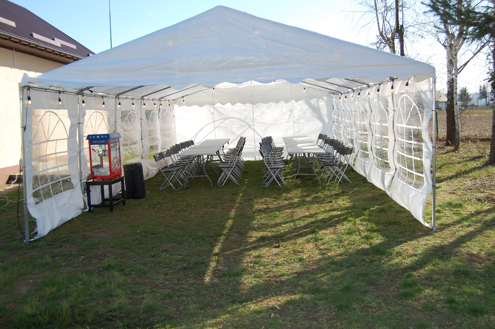Biały namiot eventowy z oknami, oświetlony girlandami, z ustawionymi stołami i krzesłami, oraz maszyną do popcornu, na trawiastym terenie.