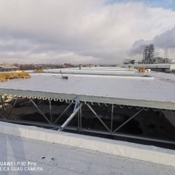 PRO-DACH Patryk Arent - Rozległy dach płaski w trakcie prac, widoczny pracownik w oddali, palety z materiałem i maszyna do papy, kominy fabryczne w tle.