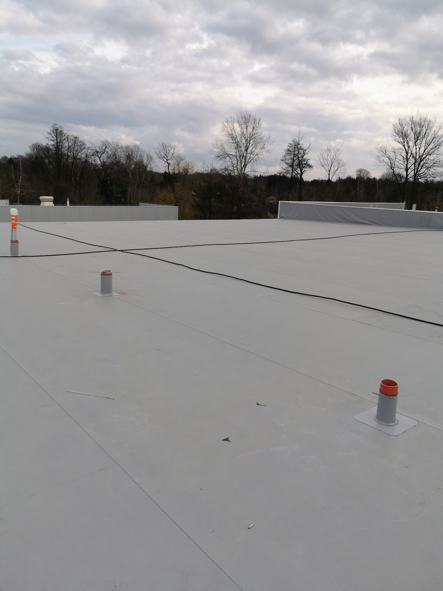 Nowe, płaskie pokrycie dachu przemysłowego w Kolbudach z widocznymi kominkami wentylacyjnymi i systemem odwadniającym, na tle pochmurnego nieba.